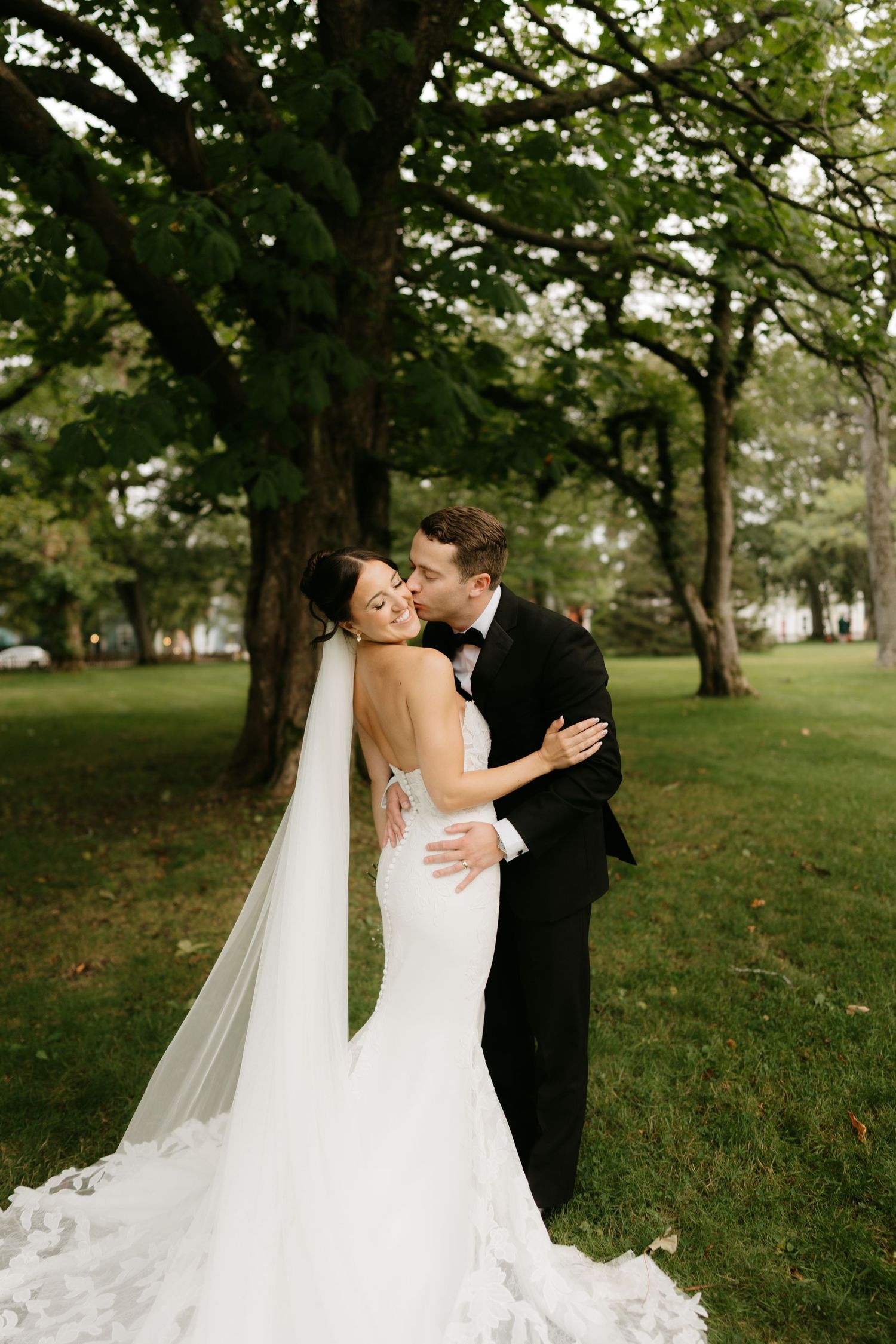 Bride in white mermaid gown and veil embraced by groom in black tuxedo, sharing a tender kiss outdoors under lush green trees.