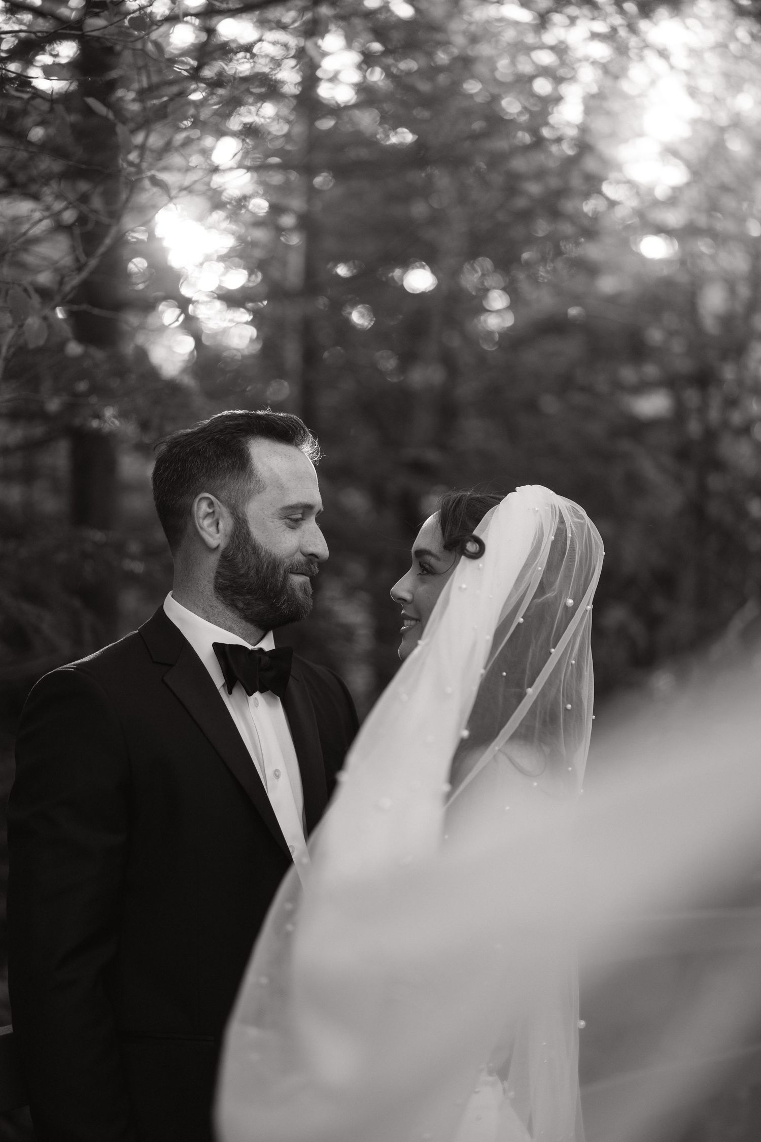 Black and white photo of a bride and groom in formal attire gazing at each other in a forest setting.