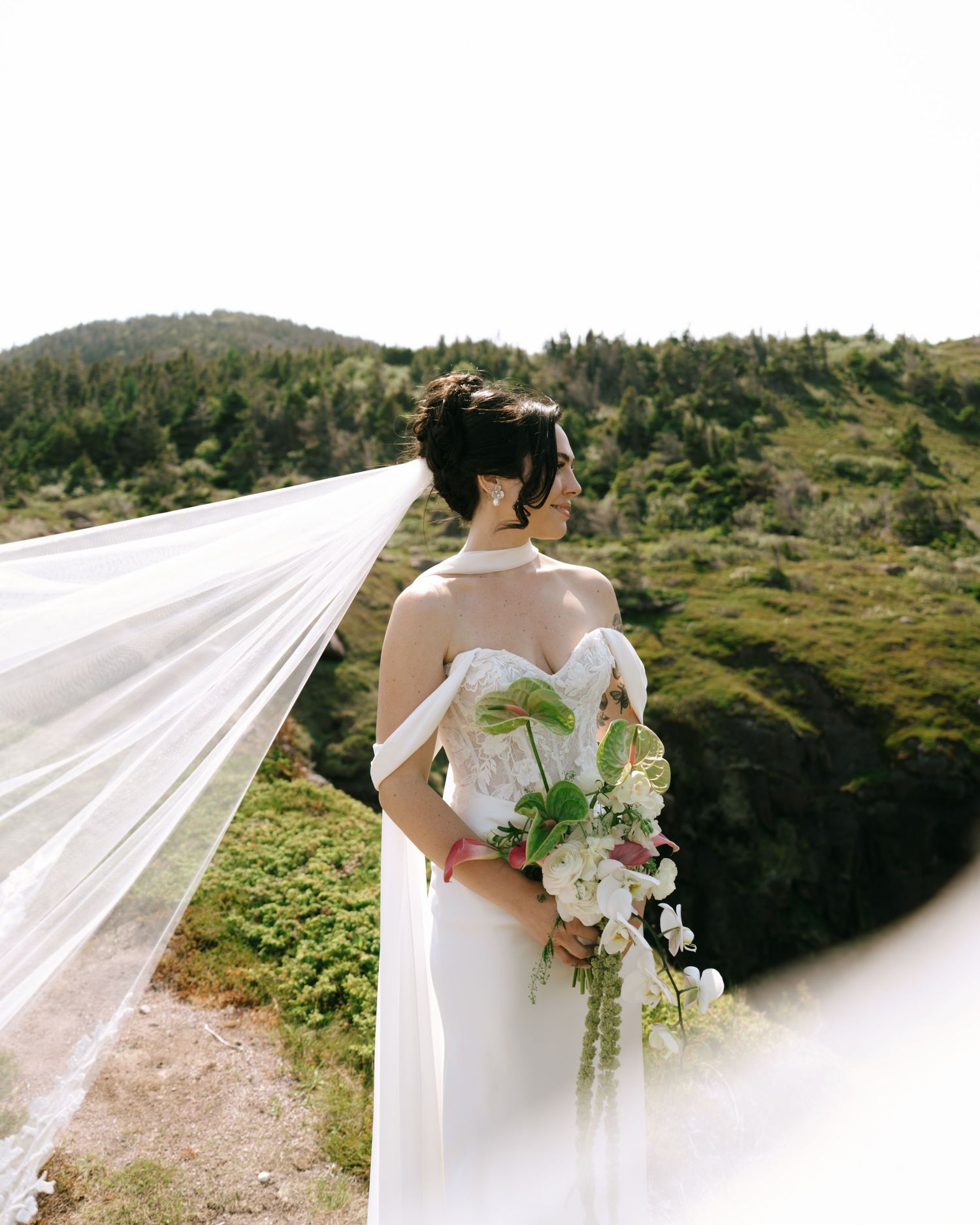 Bride in off-shoulder gown holds floral bouquet, veil flowing in wind, with lush green hills backdrop.