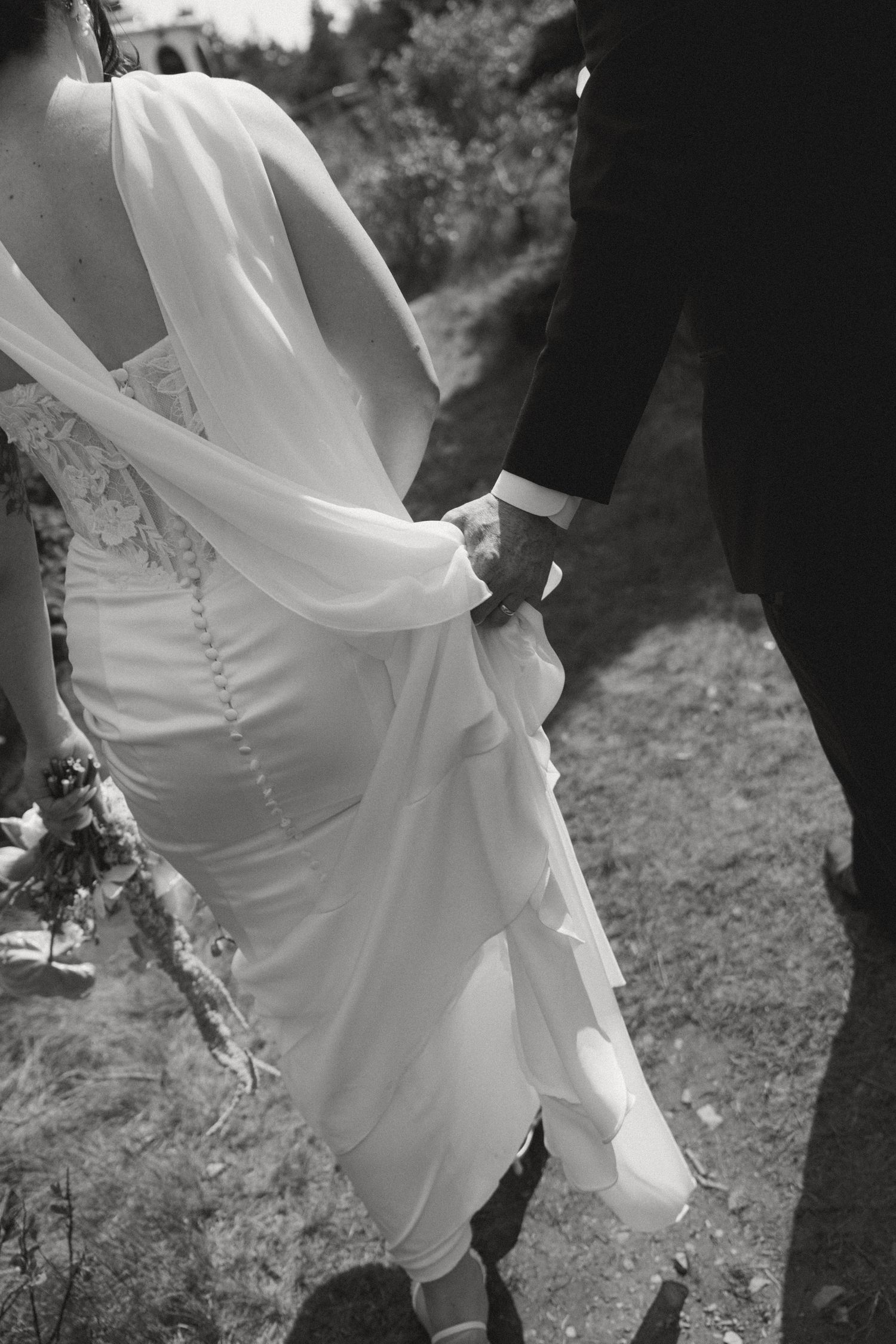 Black and white photo of bride and groom holding hands, bride wearing elegant flowing wedding dress with sheer cape.