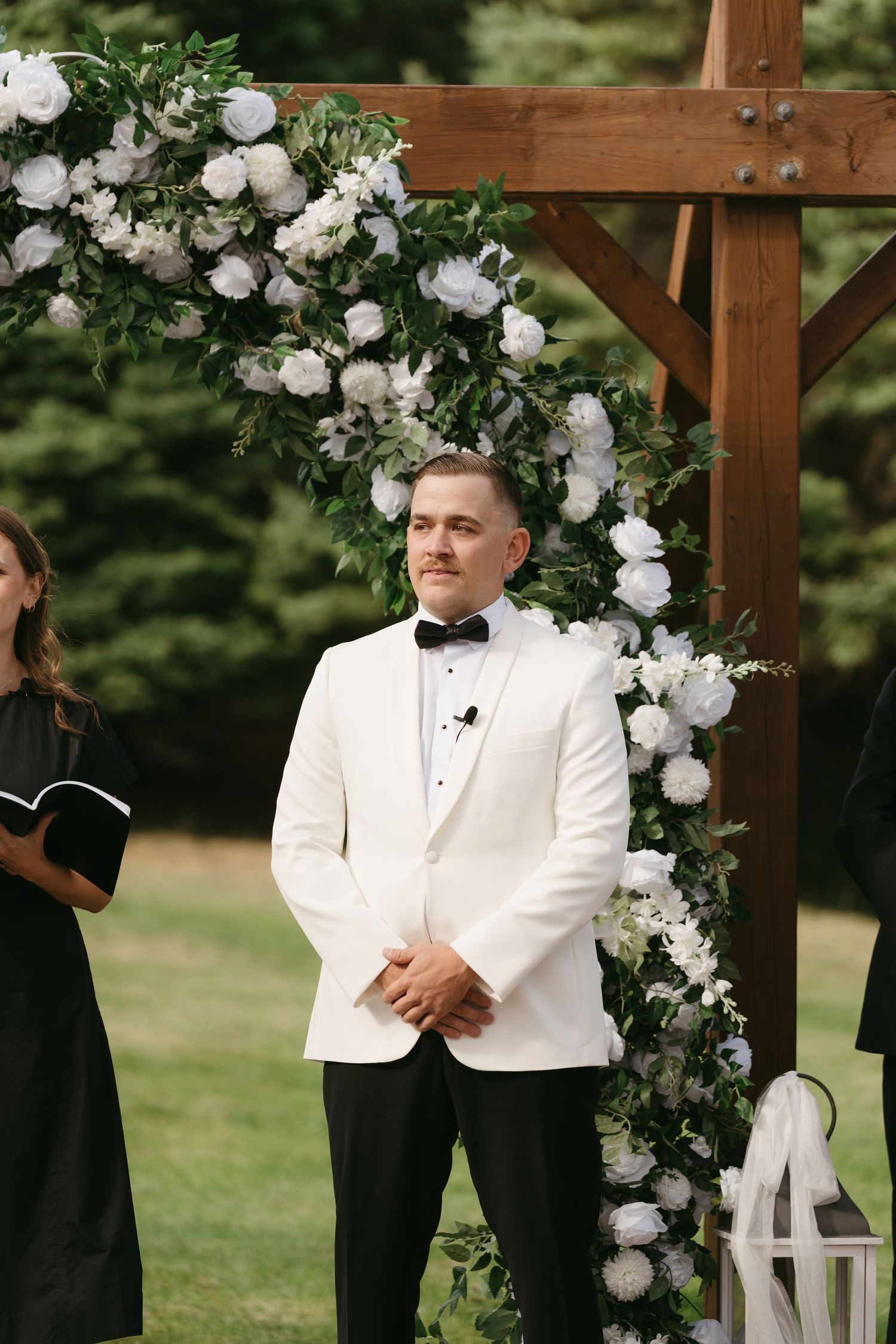Wedding ceremony setup with white floral arch decorations and people standing at an outdoor venue.