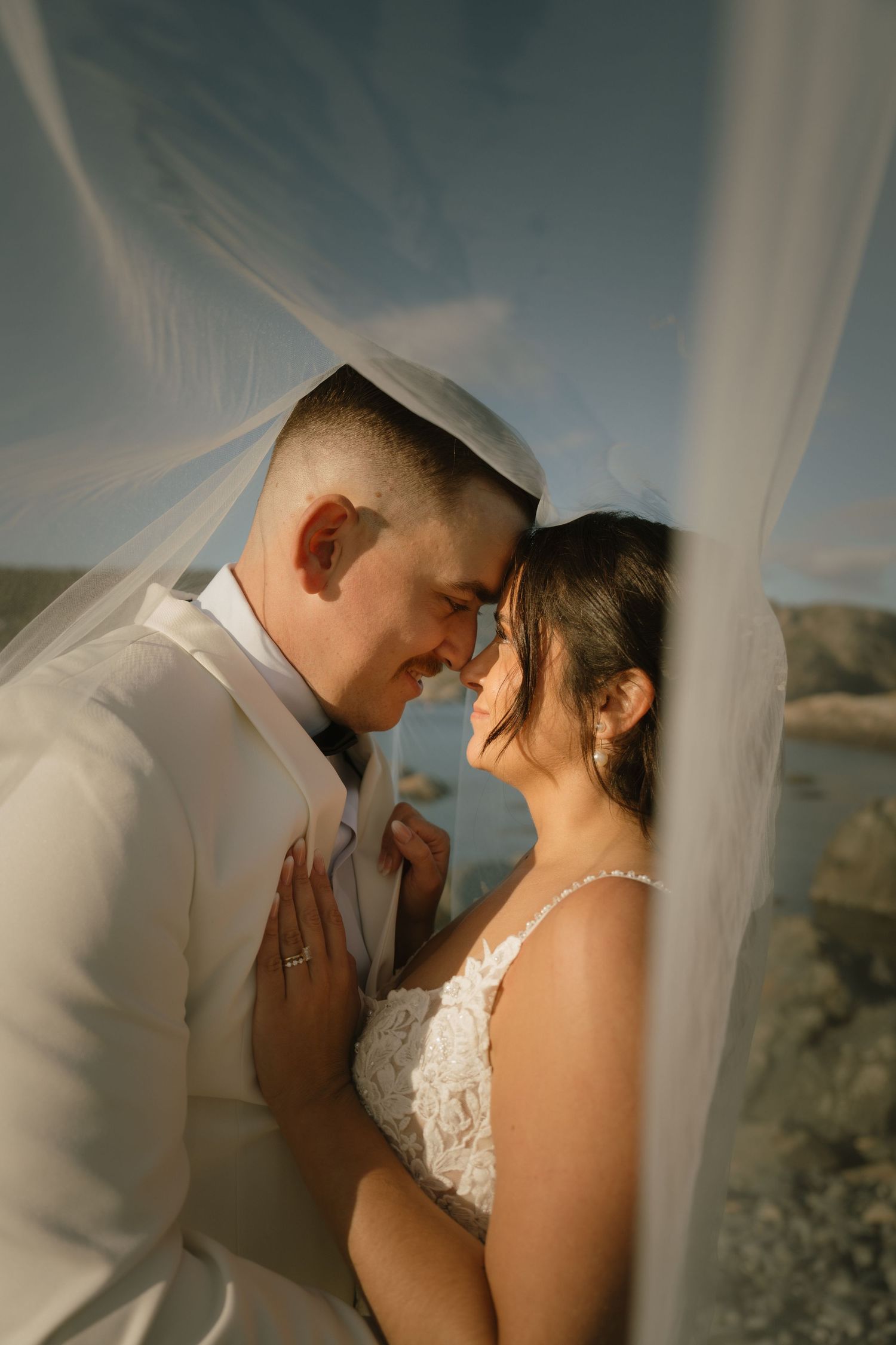 A romantic wedding couple shares an intimate moment under a flowing veil against a scenic outdoor backdrop.