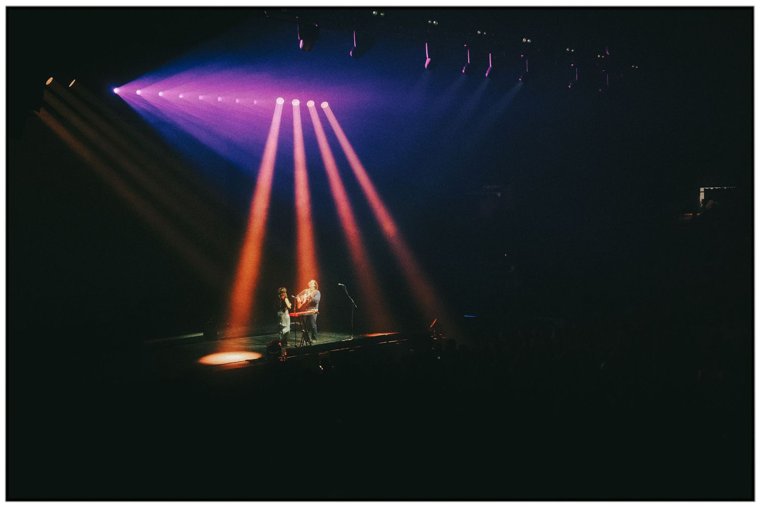 Concert stage lighting beams create dramatic purple and orange rays through atmospheric haze during a live performance.