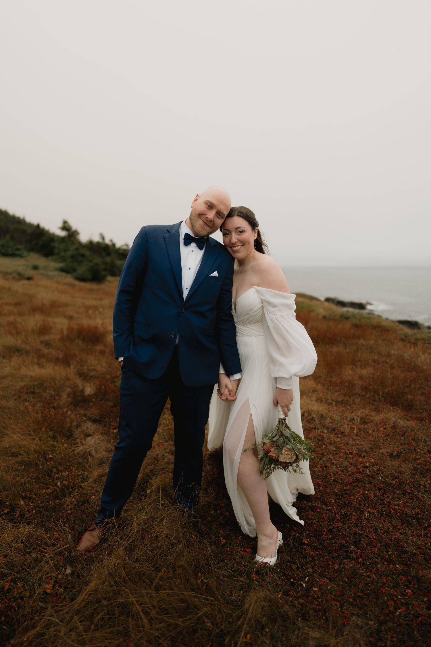 A couple poses together on a coastal hillside, wearing formal wedding attire against a moody seaside backdrop.