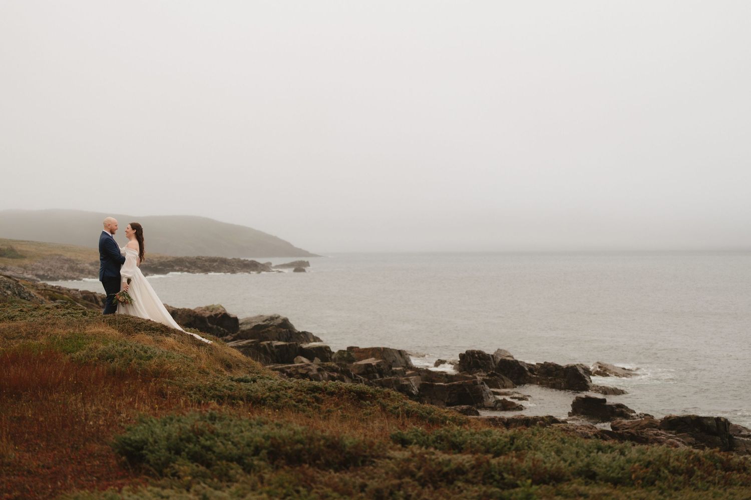 Couple stands on rocky coastal cliffs overlooking misty ocean views during outdoor wedding ceremony.