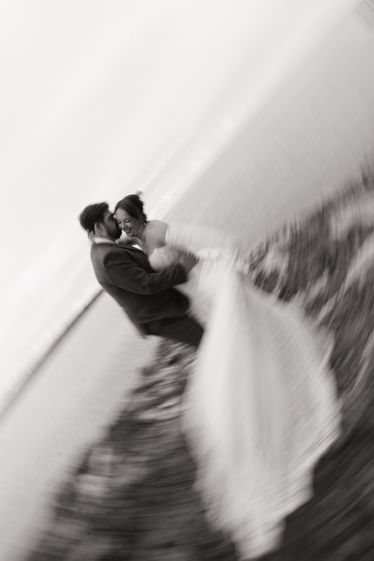 Black and white wedding photo of bride and groom embracing near water, captured with artistic motion blur effect.