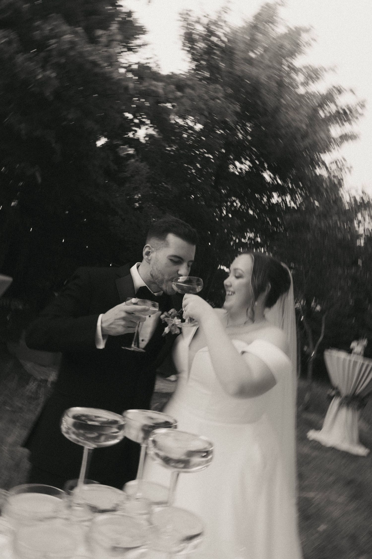 Couple in wedding attire share drinks during an outdoor celebration.