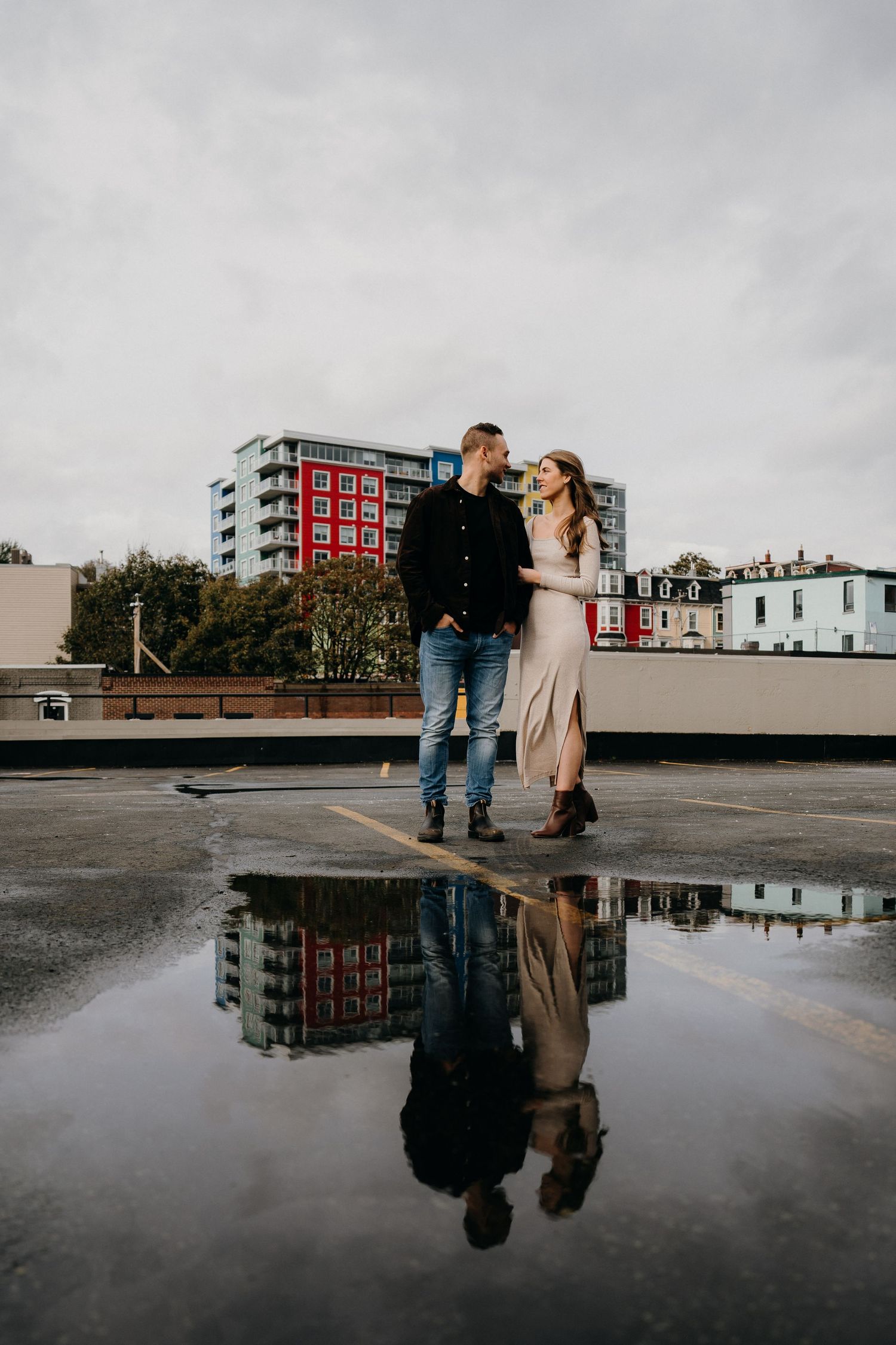 Two people embrace on a city rooftop with their reflection captured in rainwater below.