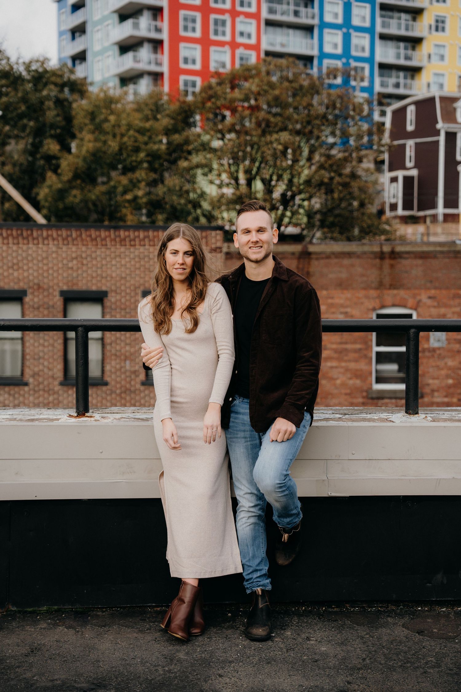 Couple standing close on rooftop parking area with brick buildings and autumn trees in background.