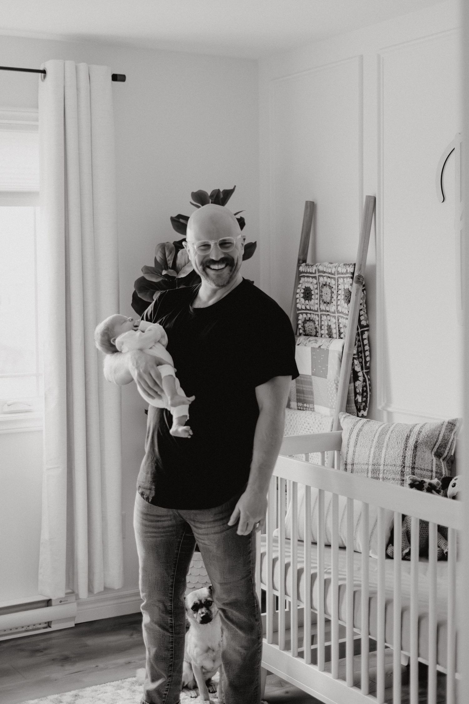 Person in casual attire holding baby while standing next to white crib in bright room.