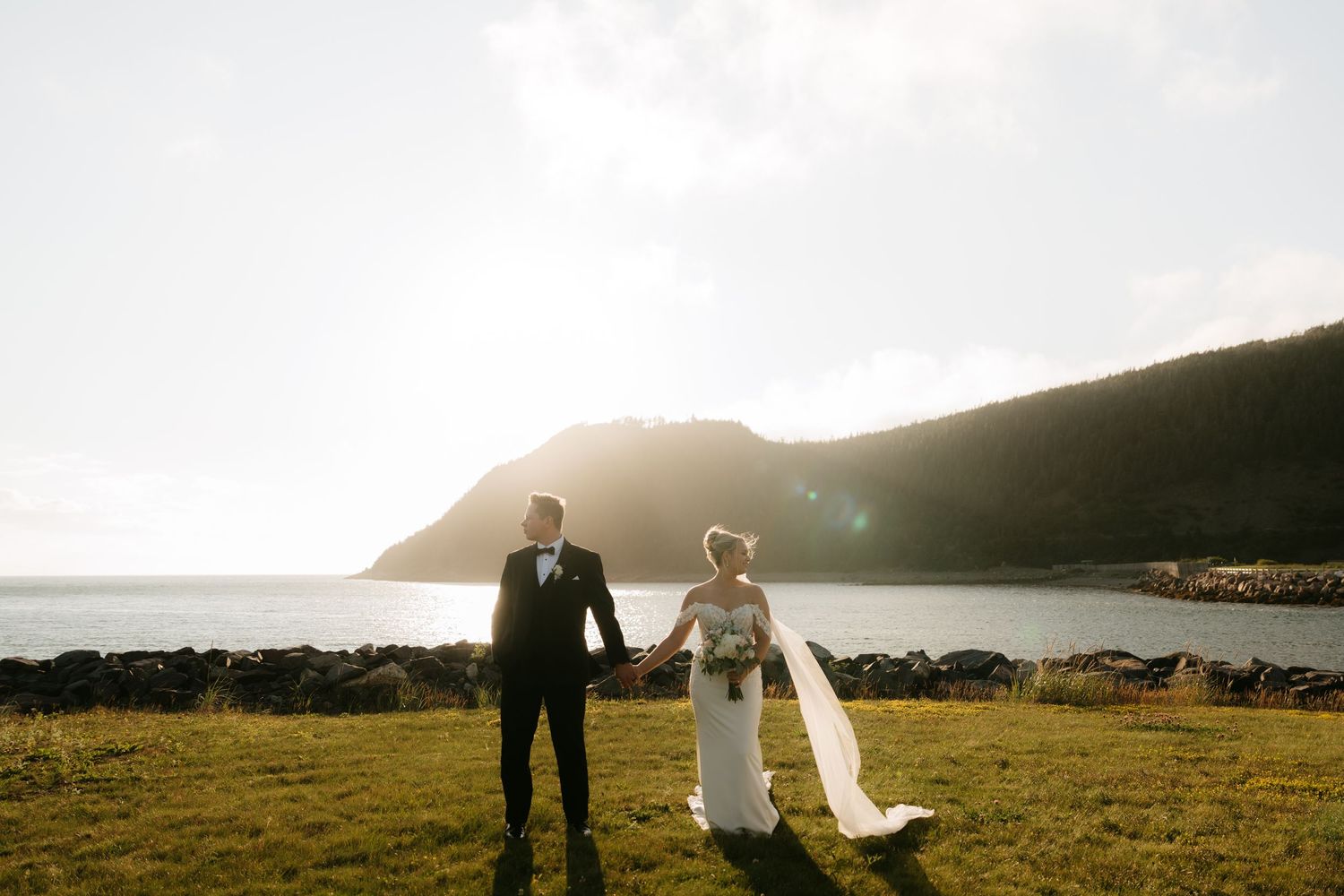 Bride and groom holding hands at golden hour overlooking a scenic coastal bay with green hills.