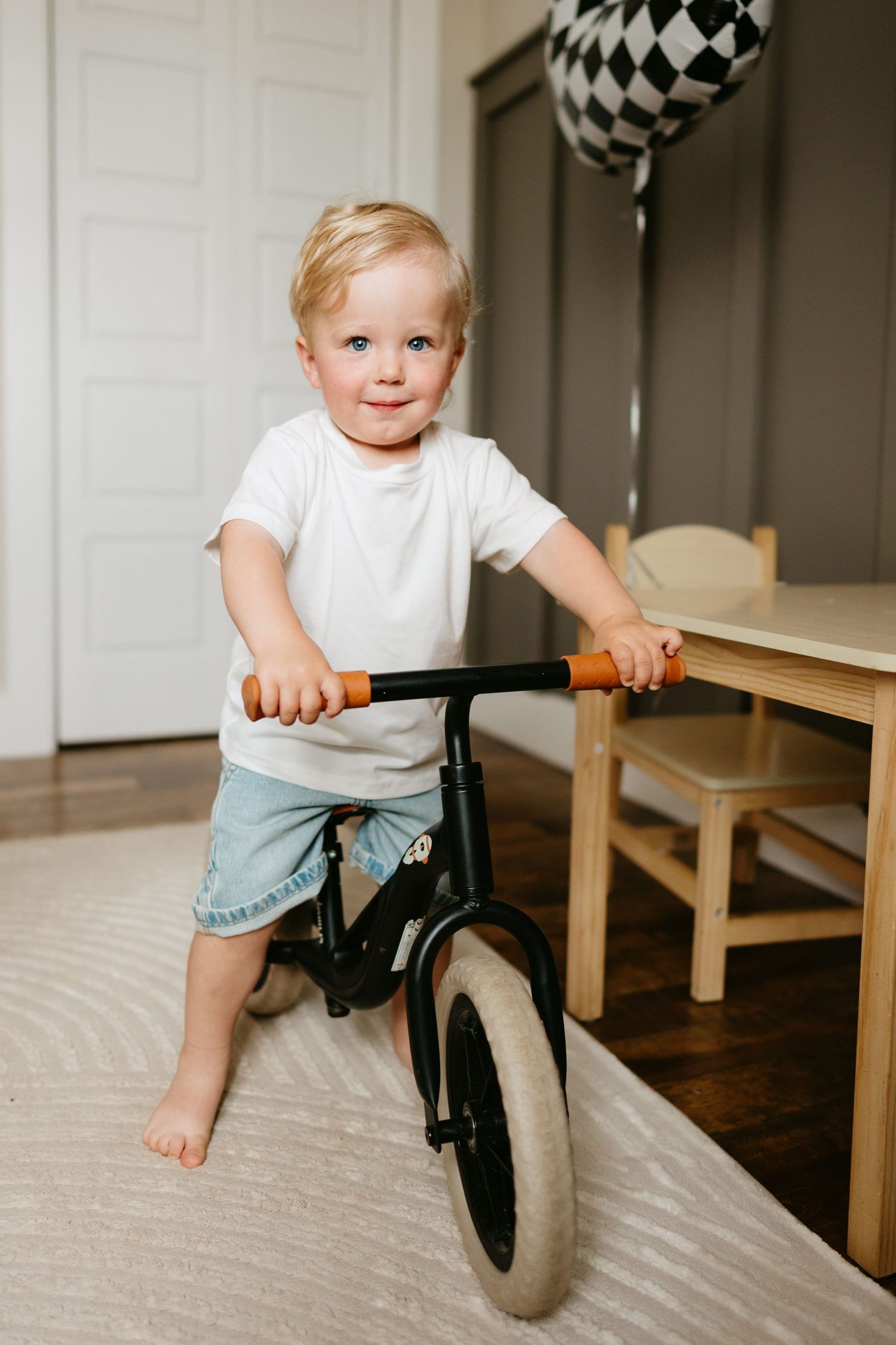 Toddler boy in white t-shirt riding a balance bike indoors on a rug near a wooden table.