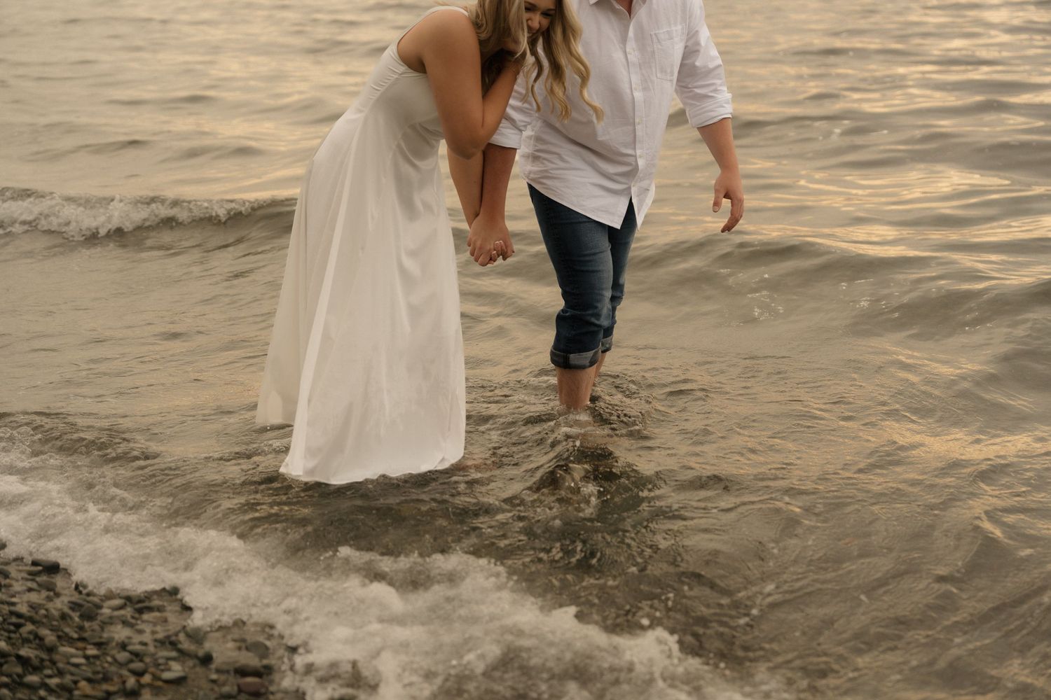 A couple in wedding attire walks along the beach shoreline at sunset with waves lapping at their feet.
