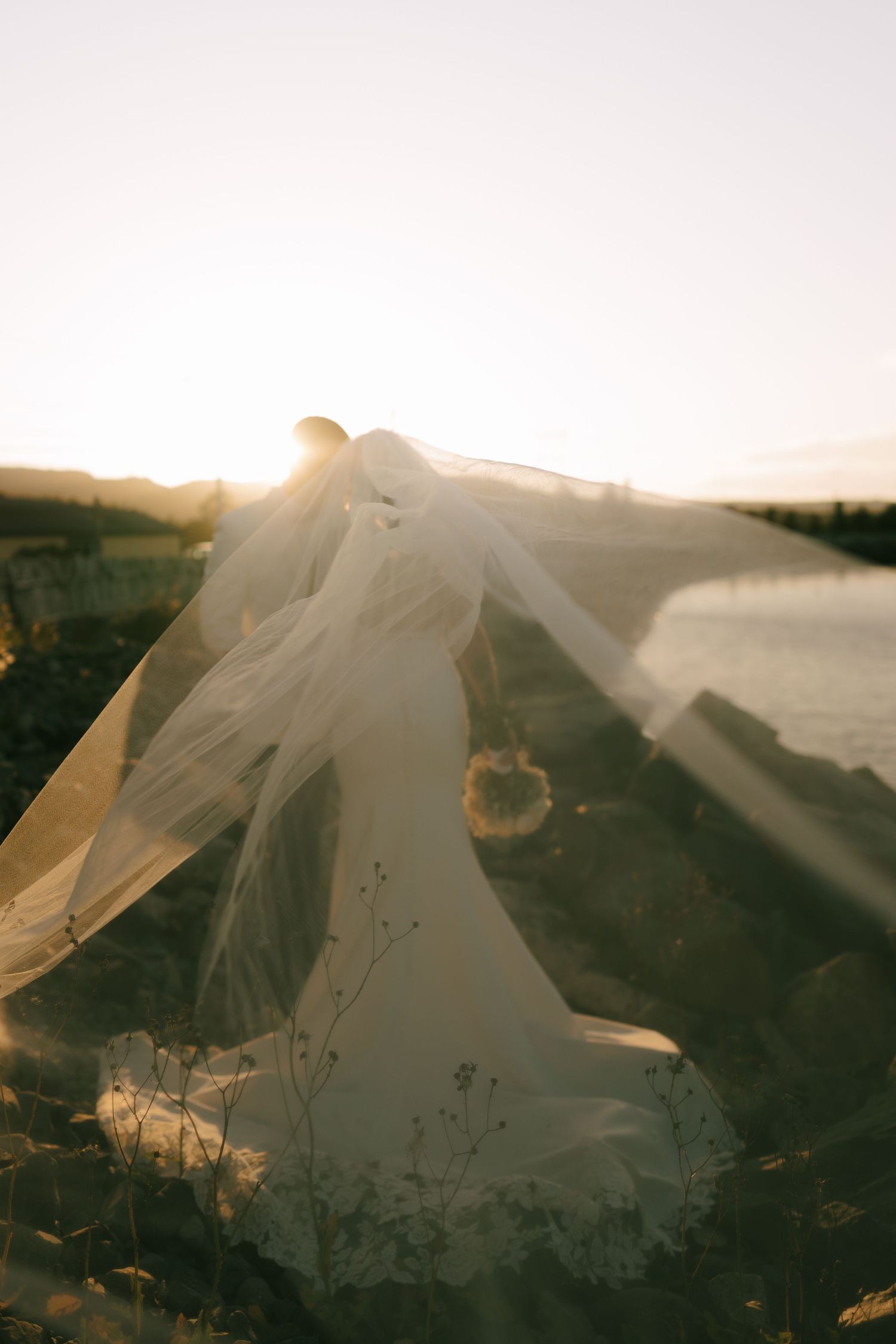 A flowing white wedding veil catches sunlight and billows in the breeze against a sunset landscape.