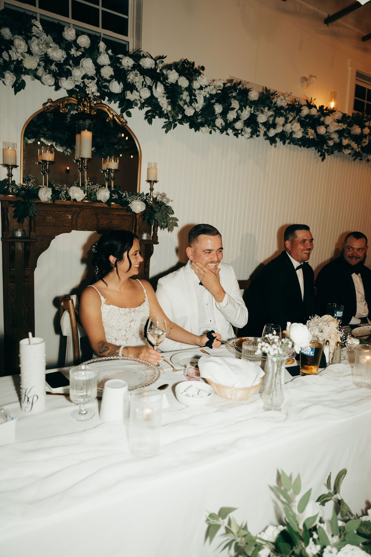 Wedding guests seated at a long banquet table with white linens and decorative greenery overhead at a reception dinner.