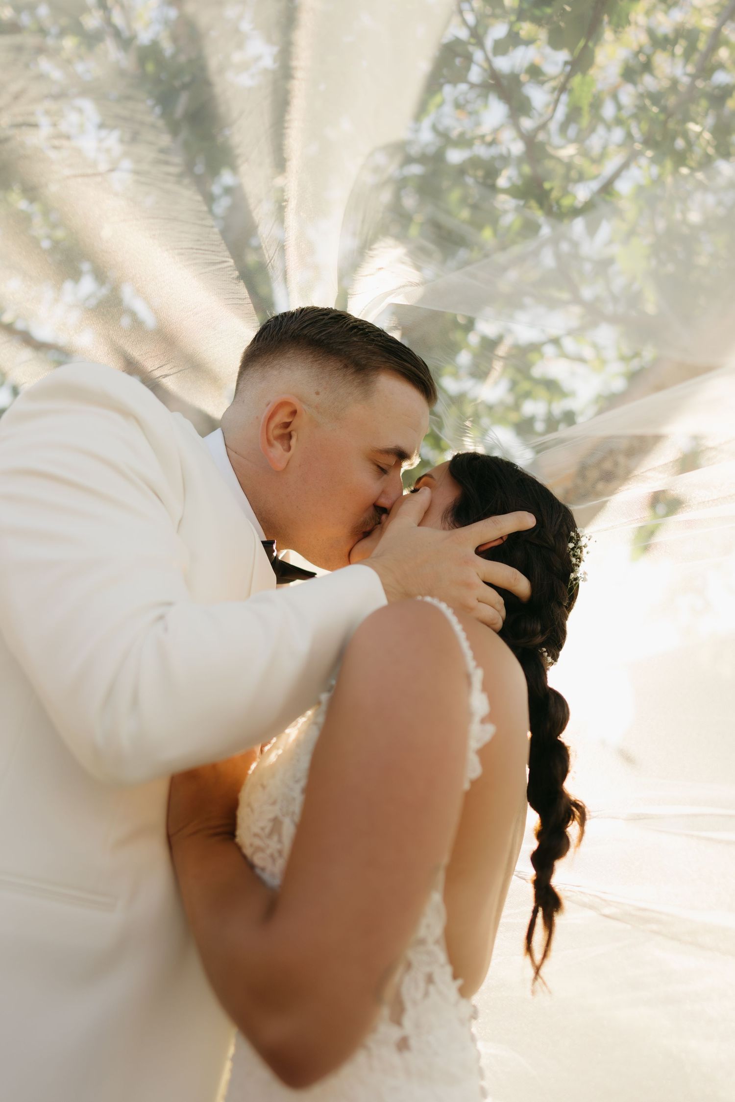 A romantic wedding moment captured in soft sunlight as a couple shares a tender kiss under a dreamy, glowing canopy.