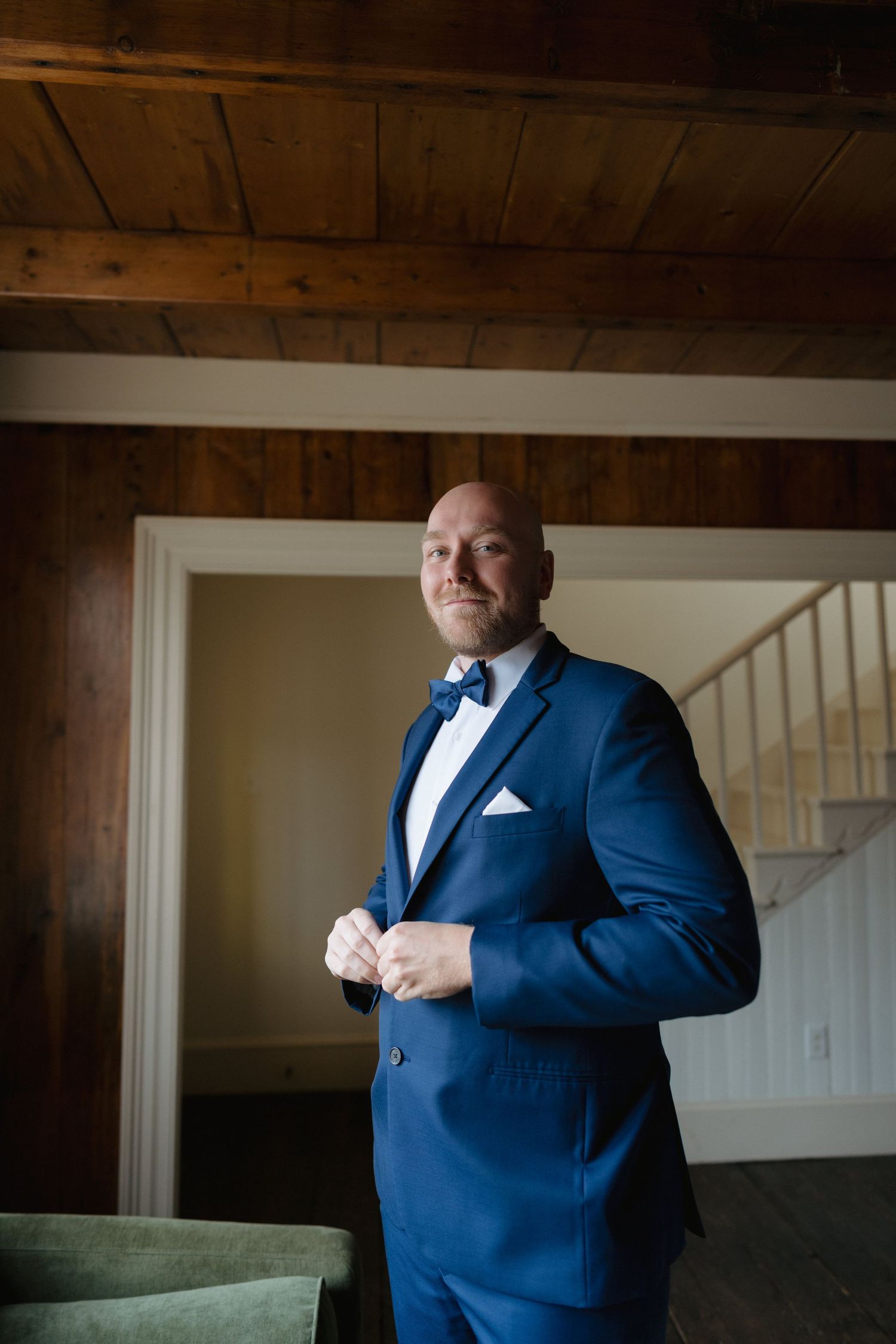 A person wearing a navy blue suit with a bow tie stands confidently in a room with wooden ceiling beams.