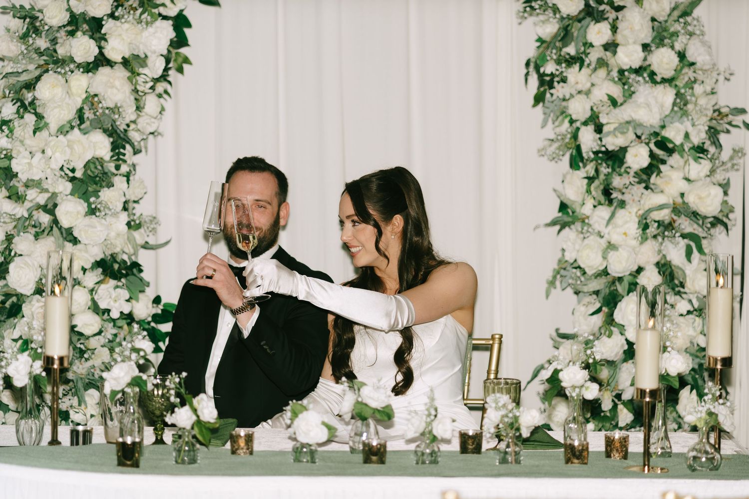 Bride and groom toast at sweetheart table adorned with white floral arrangements and candles at elegant wedding reception.