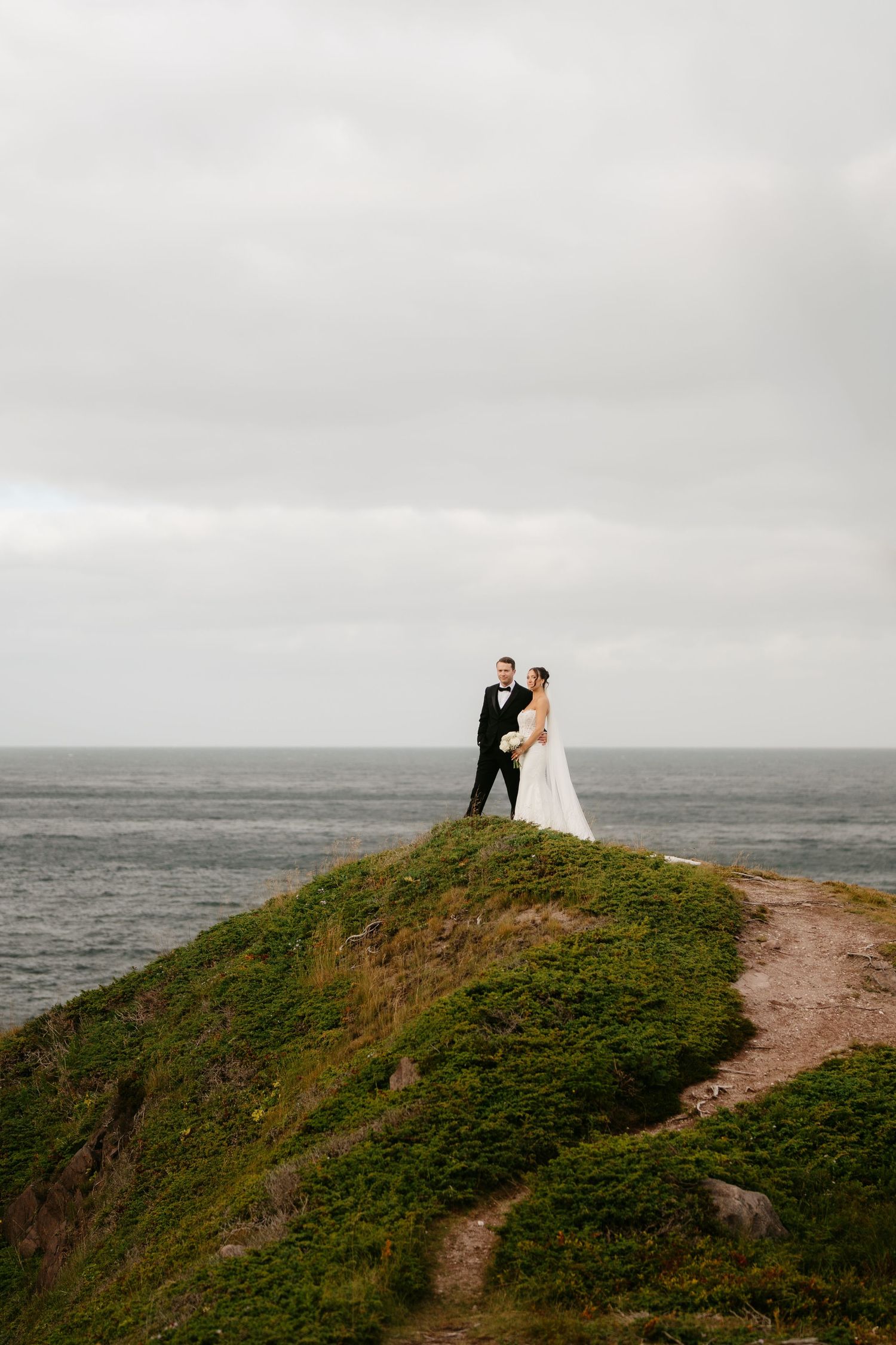 Bride and groom standing on a grassy coastal cliff with ocean views on their wedding day.