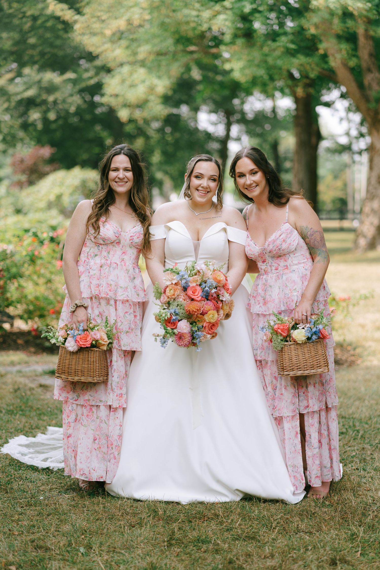 Bridesmaids in pink floral dresses holding vibrant coral and orange bouquets pose together on grassy lawn.
