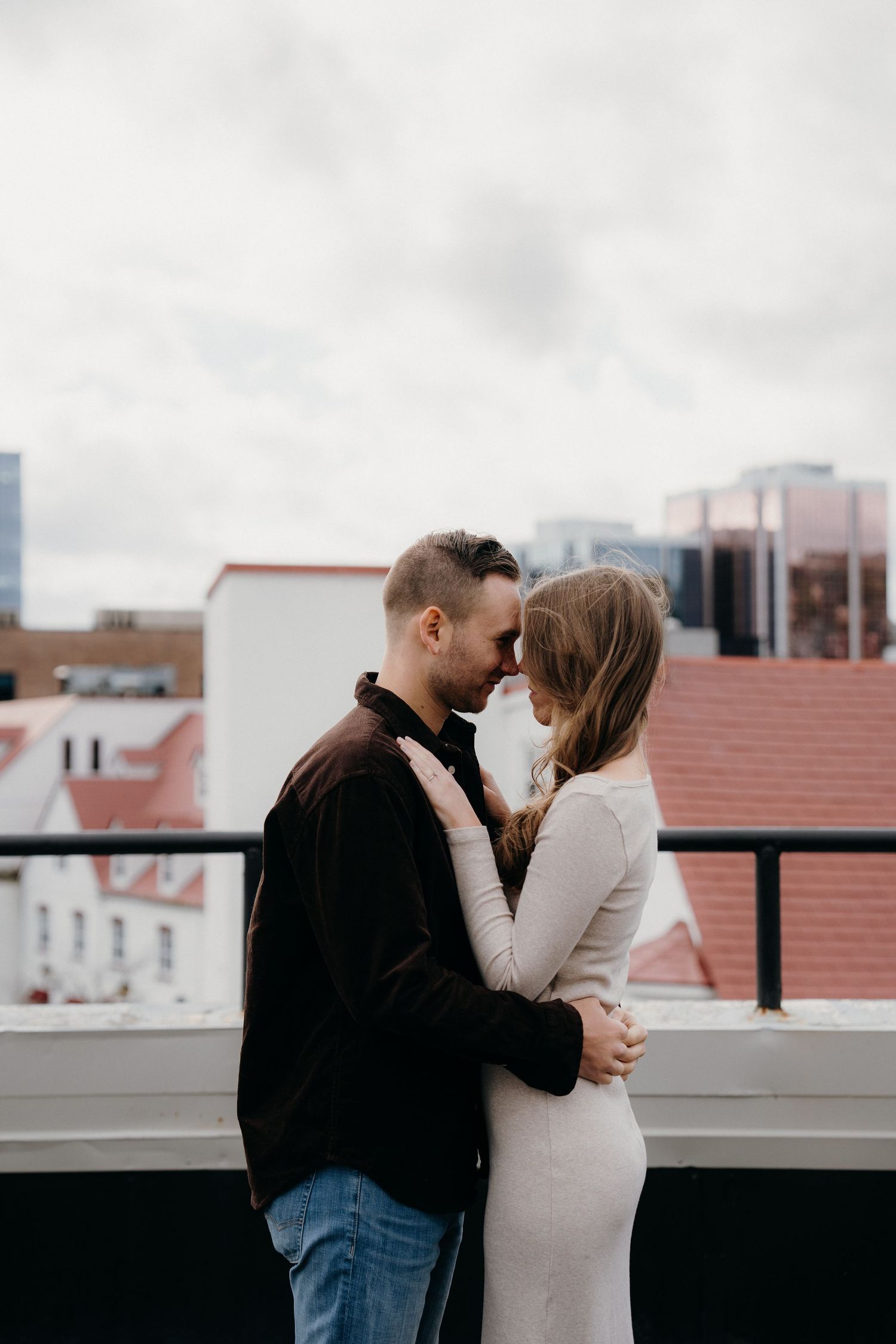 A romantic couple shares an intimate moment on a rooftop parking deck with city buildings in the background.
