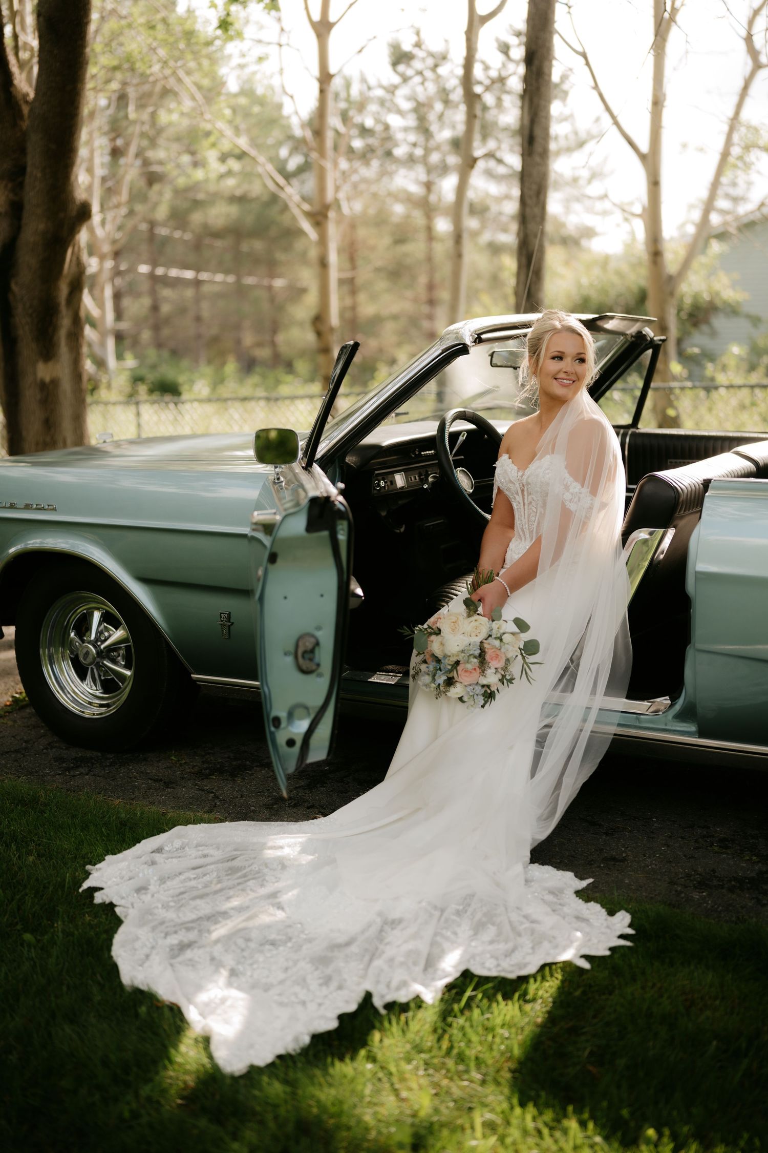 Bride in white lace gown with veil holds bouquet beside vintage teal convertible car outdoors.