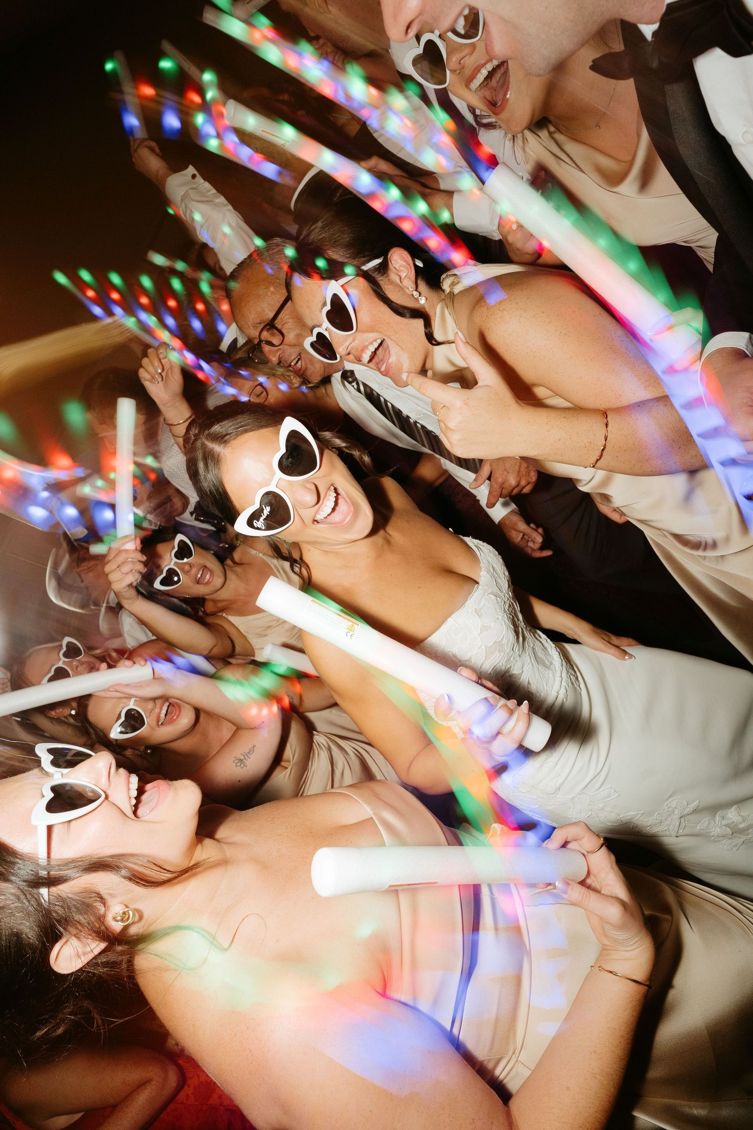 Group of party guests wearing white sunglasses holding glowing LED foam sticks at a festive celebration.