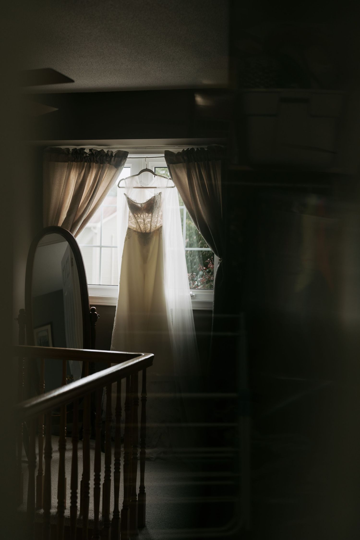 A wedding dress hanging in a window with soft natural light, draped curtains, and a staircase railing in the foreground.