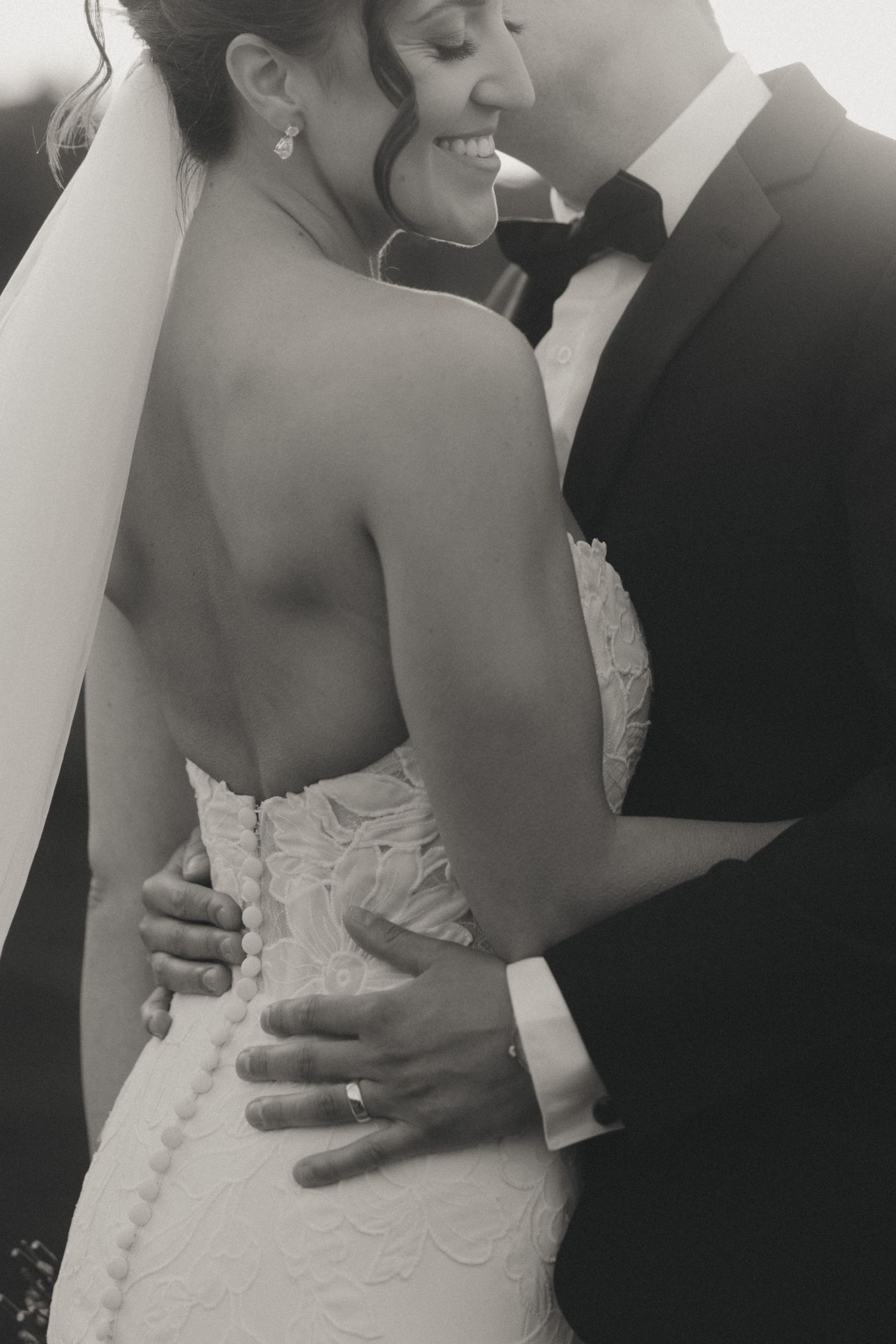 Black and white wedding photo of bride in lace gown and groom in tuxedo embracing, showing ring and veil detail.