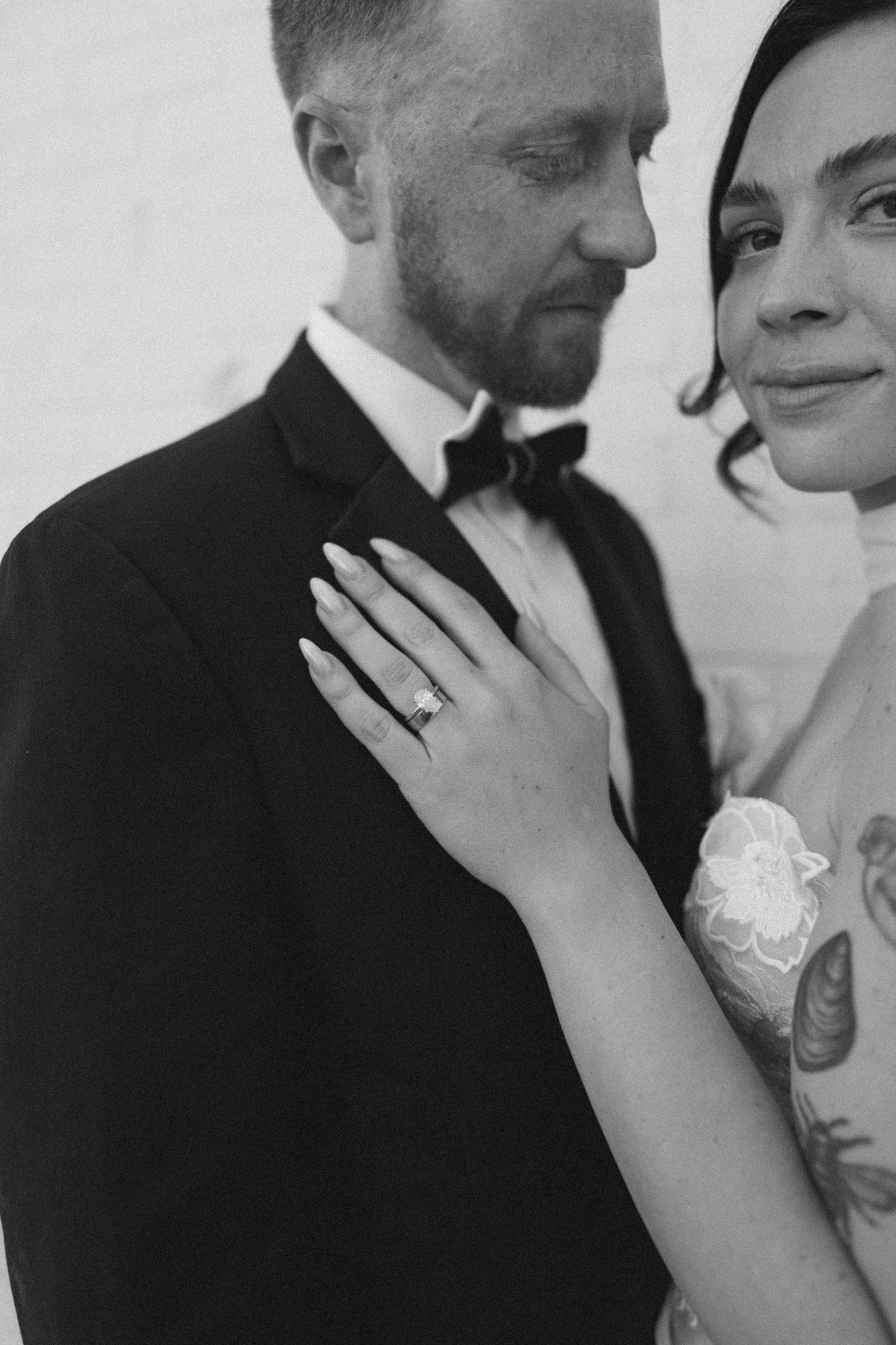 Black and white photo of bride showing diamond ring resting hand on grooms tuxedo lapel.