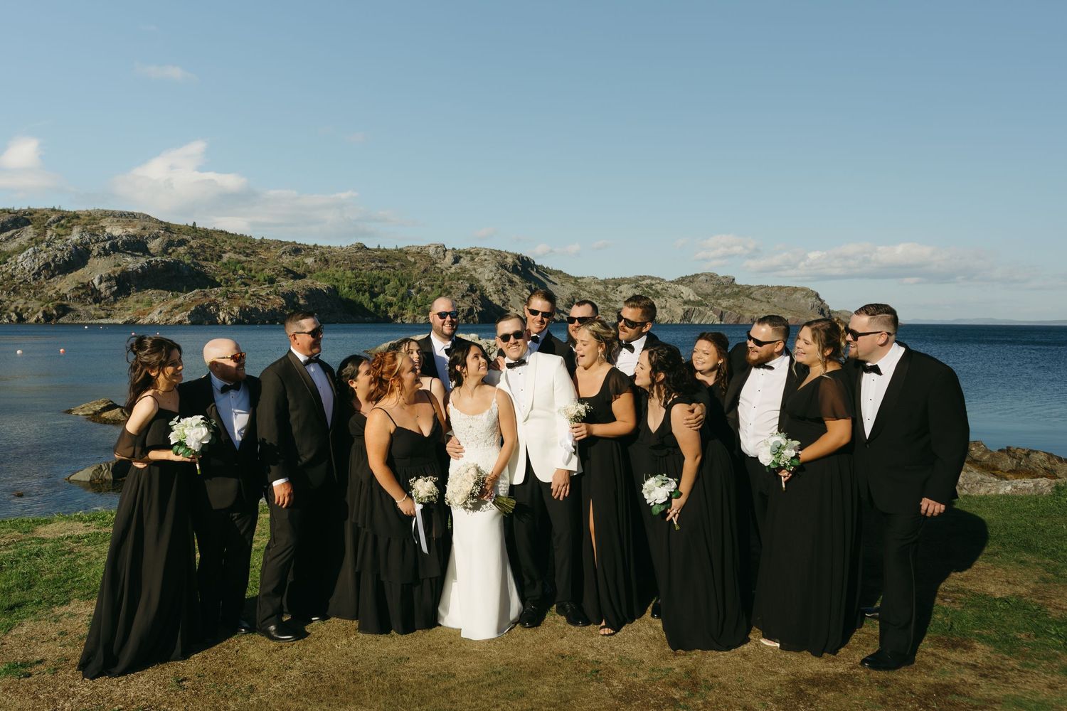 Wedding party poses together on scenic coastal cliff with ocean and blue sky in background.