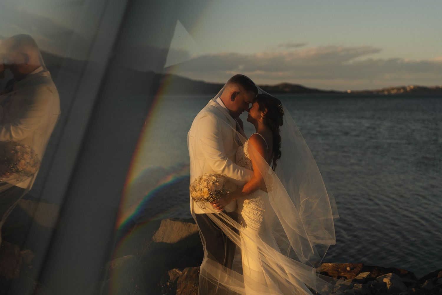 Romantic couple embraces at sunset by the ocean with mountains in the background.