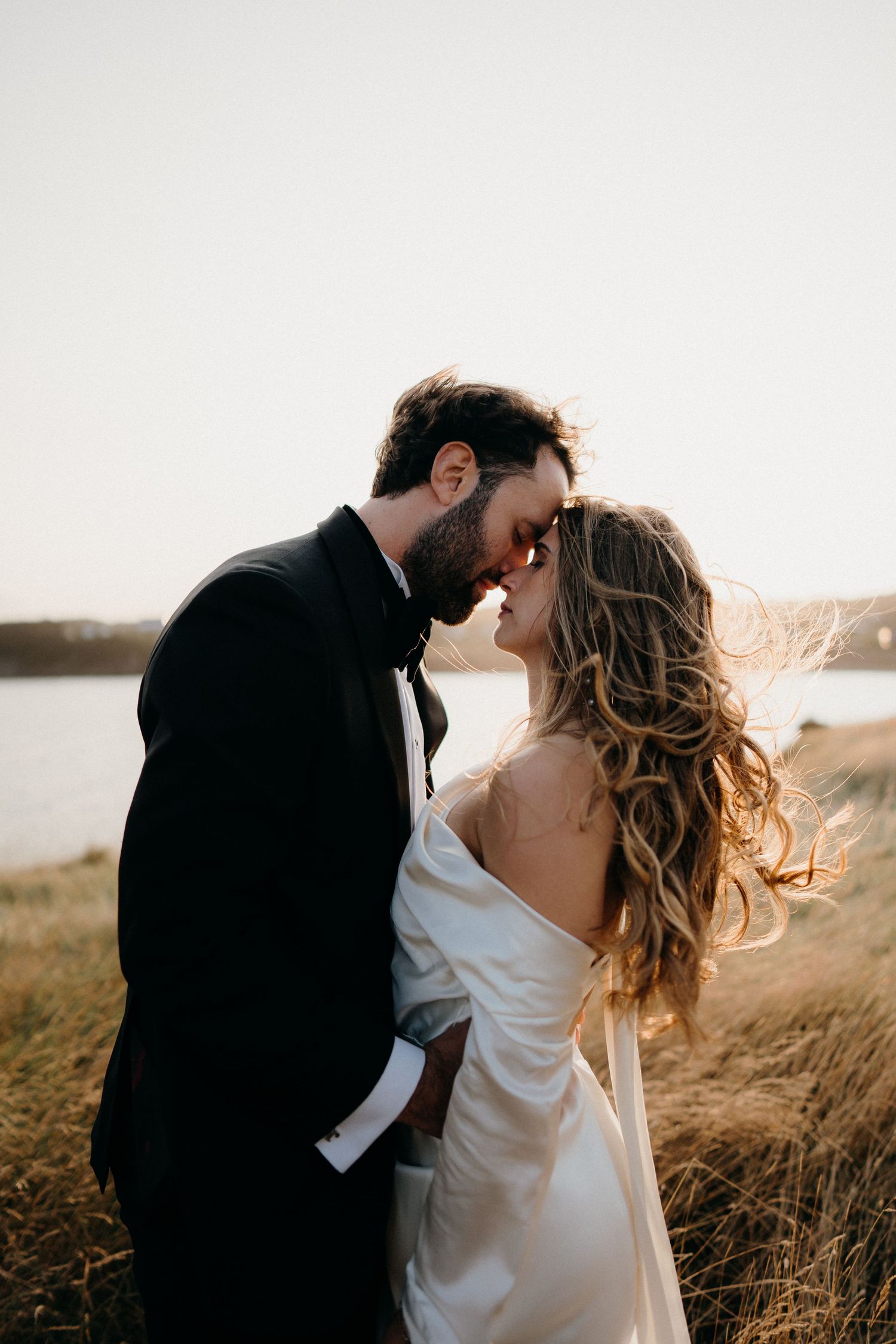 Romantic couple in wedding attire embrace at sunset on a grassy hilltop overlooking water.