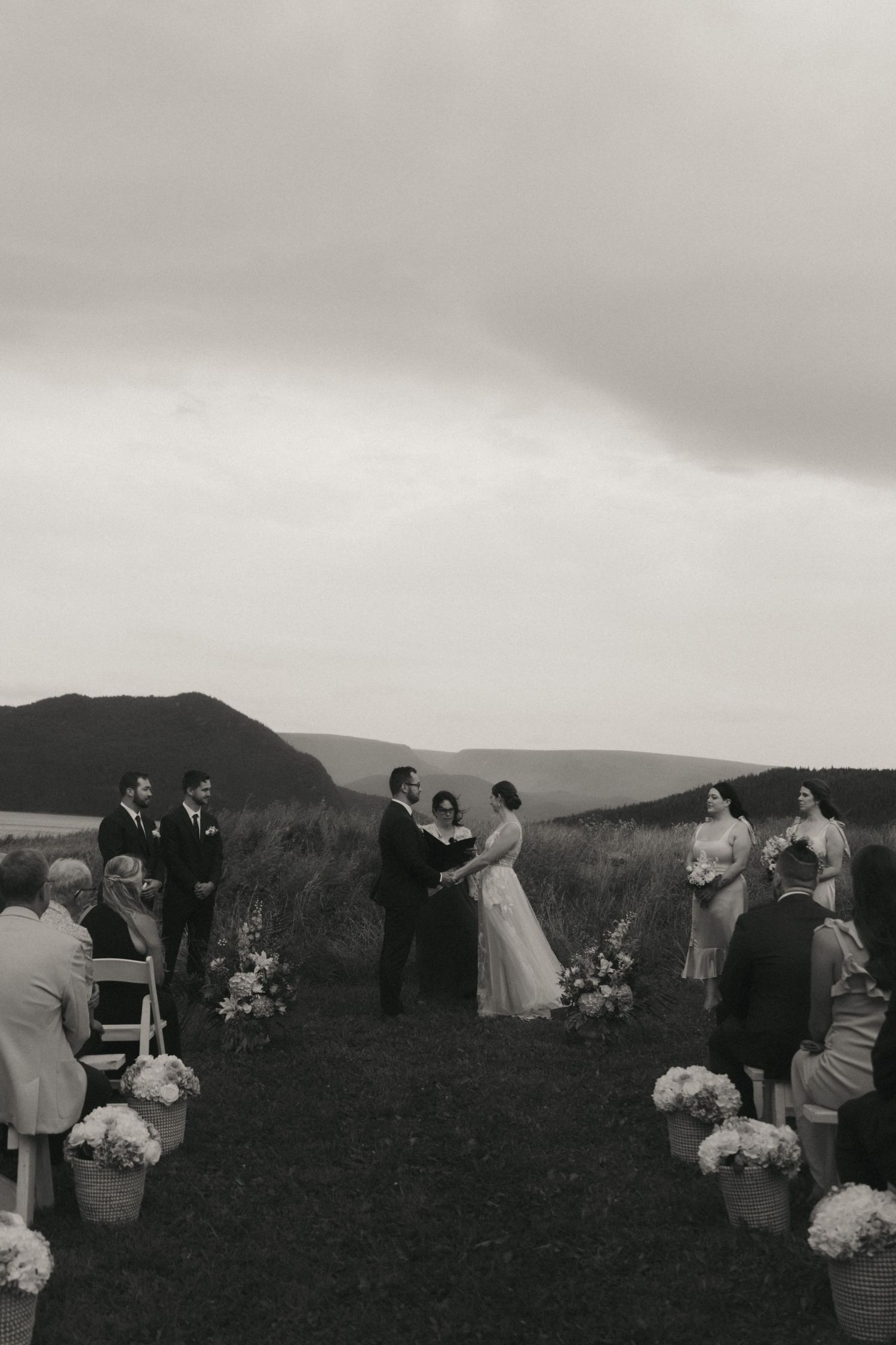 A panoramic view of a wedding ceremony taking place on a hilltop with mountains in the background at sunset.