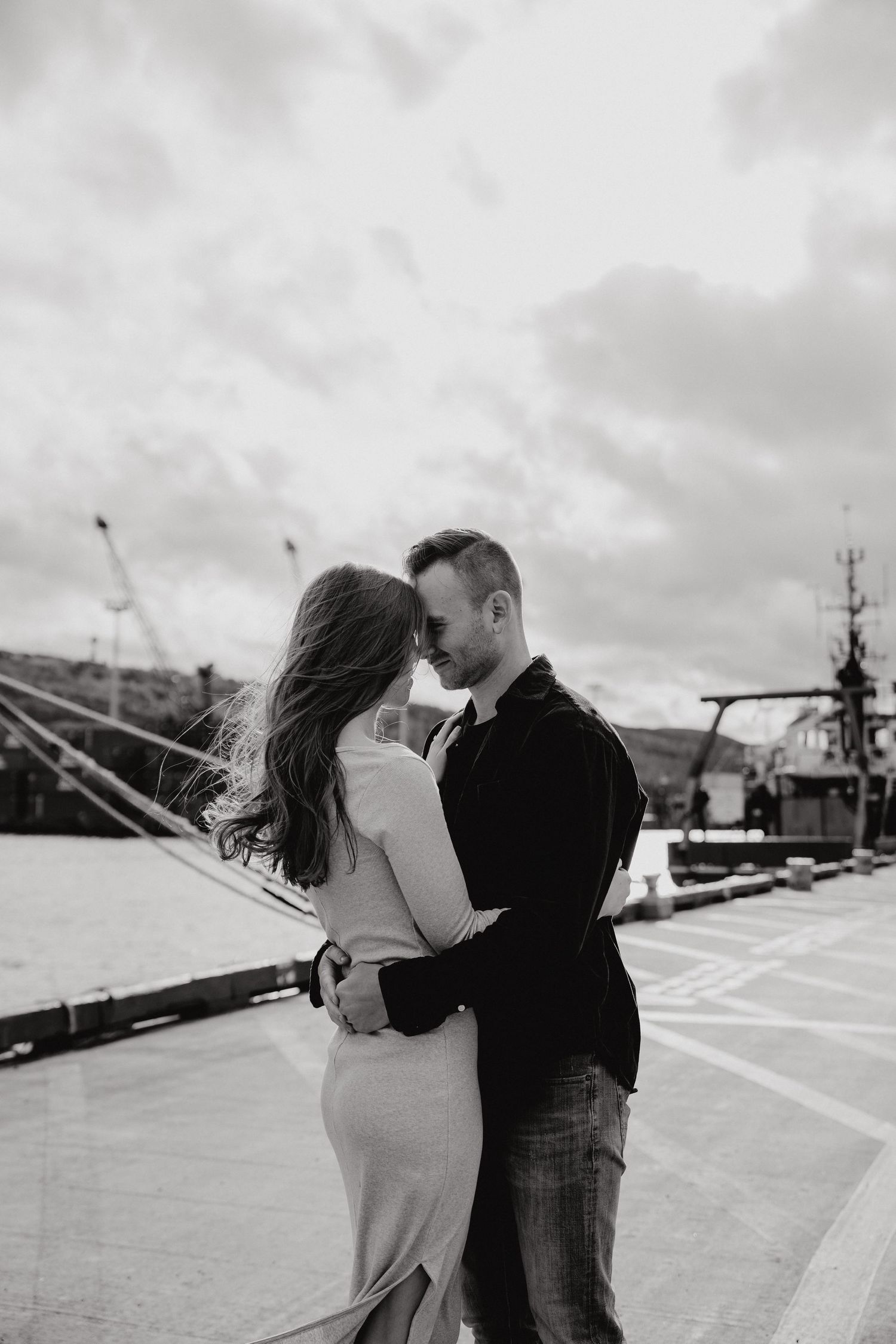 Black and white photo of couple in romantic pose against maritime industrial backdrop.