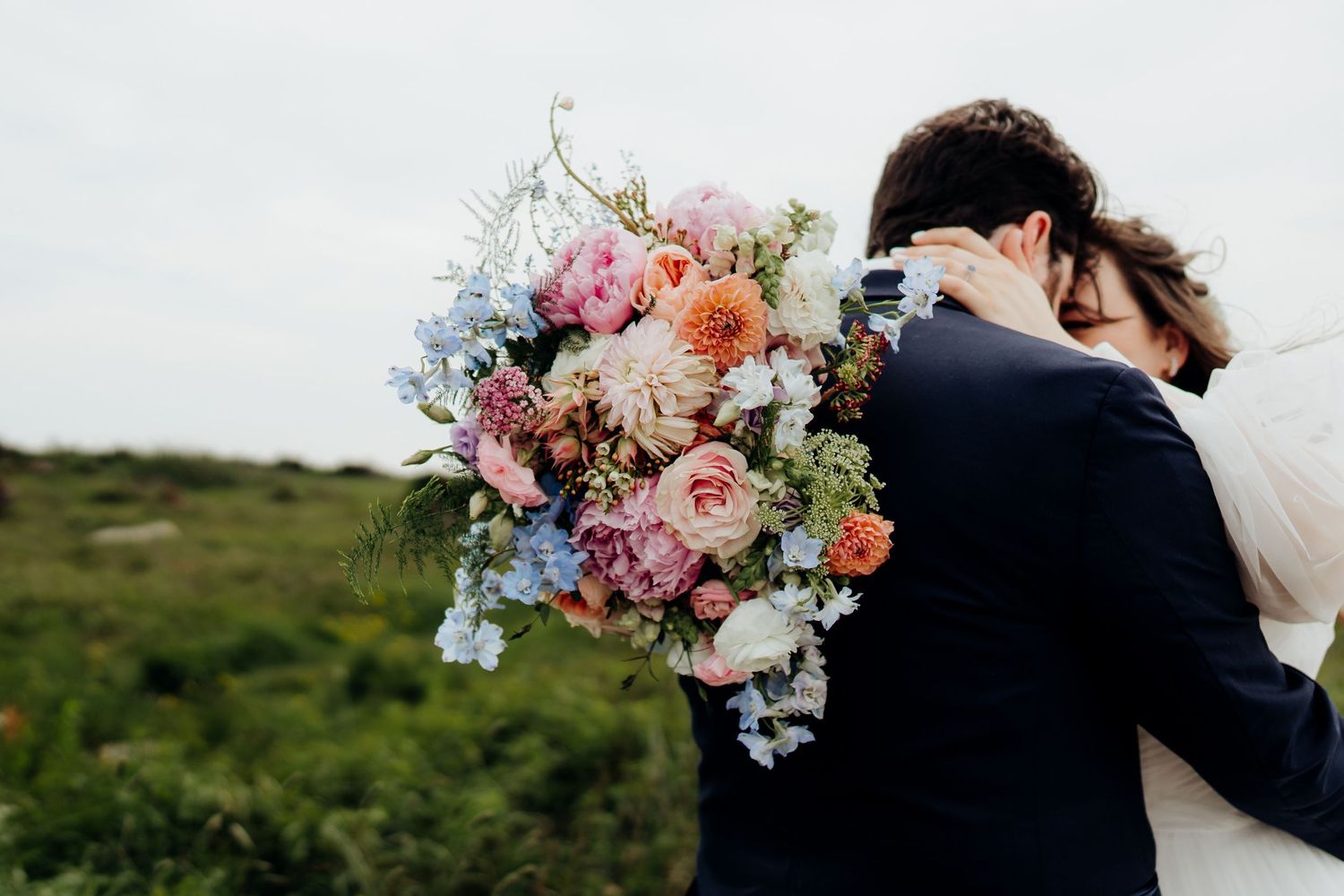 Wedding couple embraces while holding vibrant floral bouquet against natural outdoor backdrop.