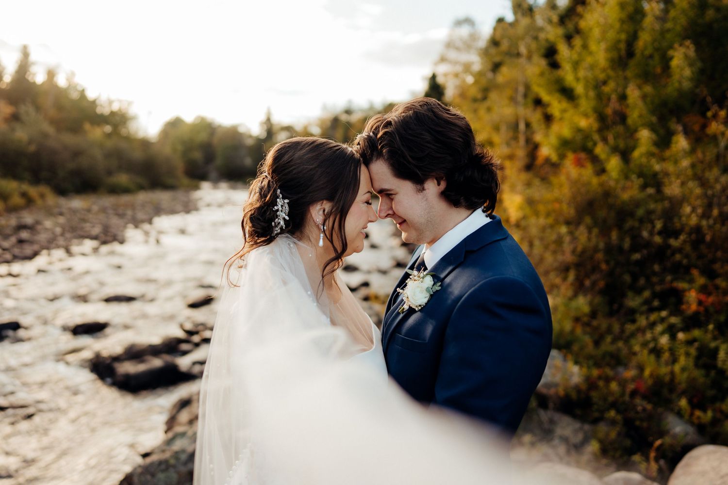 A romantic wedding moment captured at a rocky riverside surrounded by fall foliage.