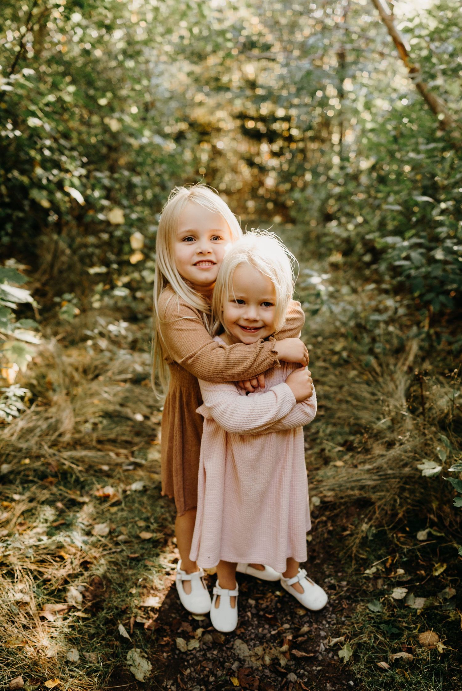 Two children in matching dresses share a sweet hug in a wooded setting.