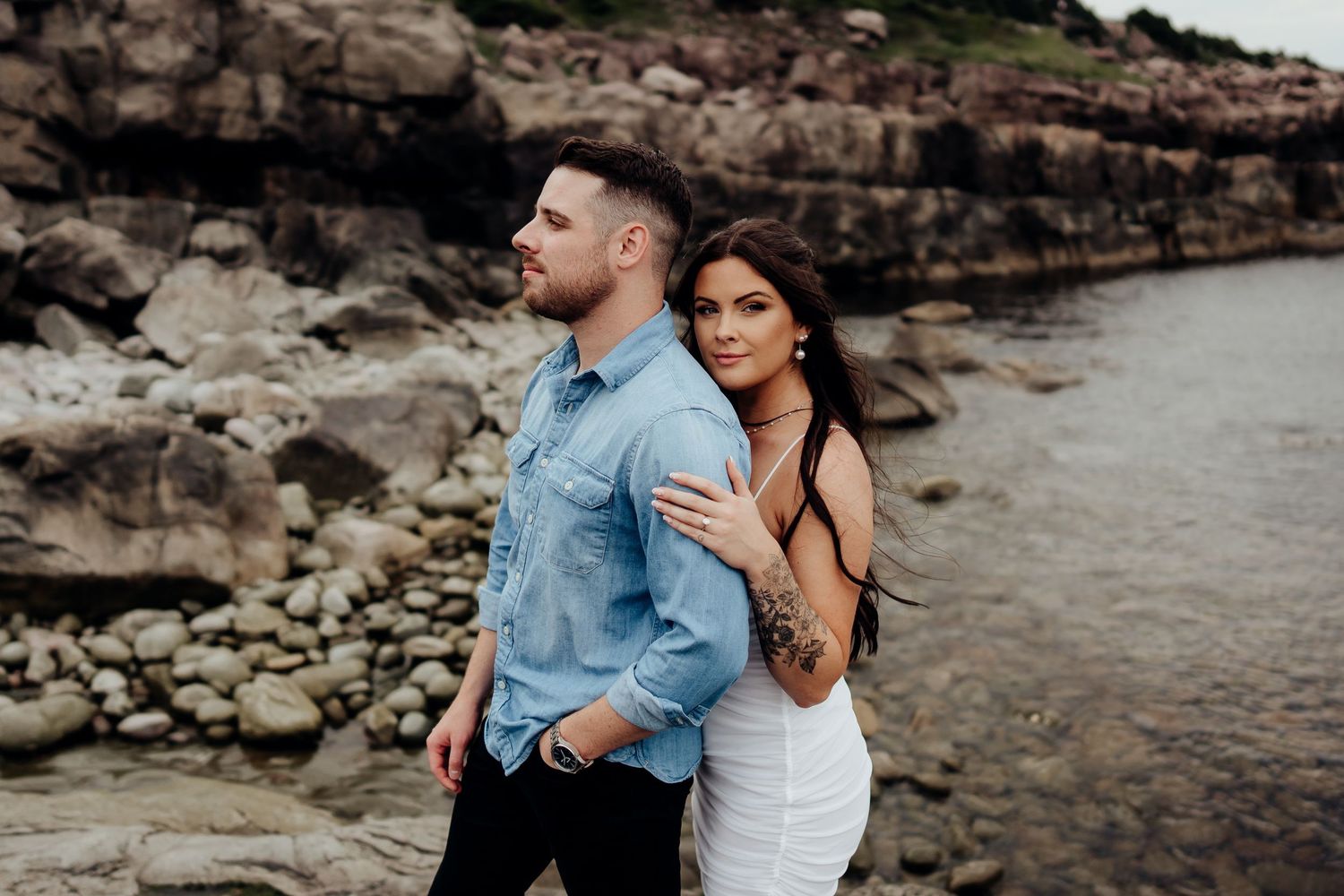 Romantic couple embraces while standing by rocky coastal cliffs at sunset.