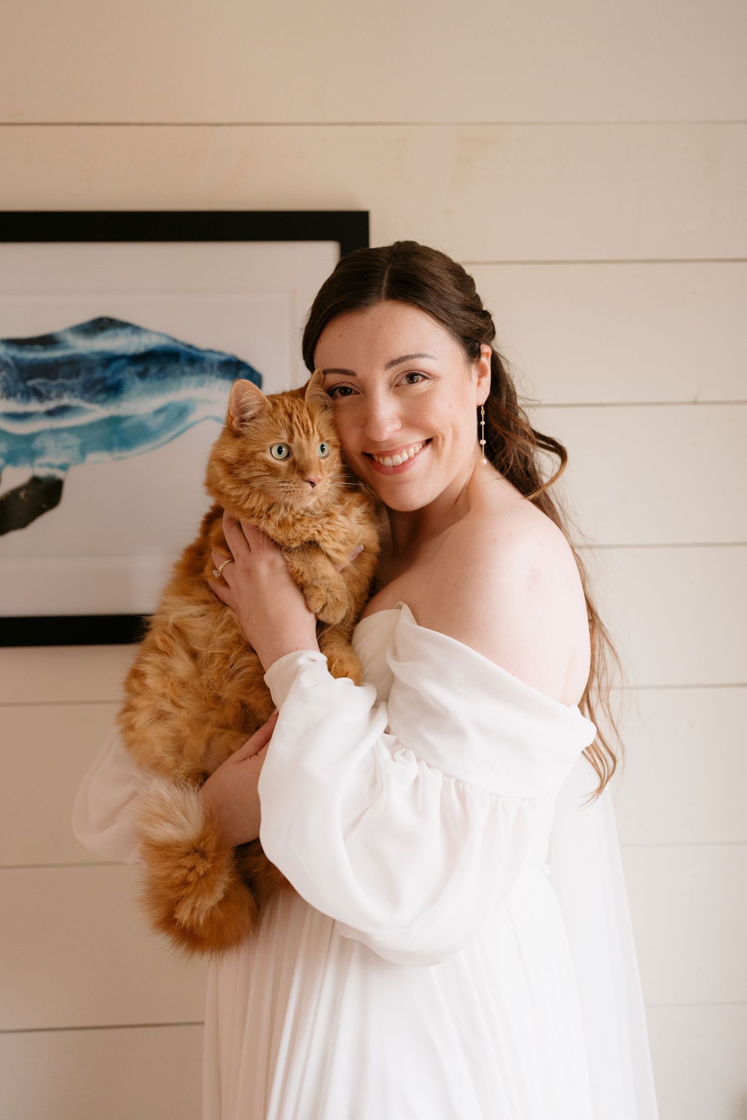 A woman in a white dress holds and cuddles with a fluffy orange cat against a light colored wall.