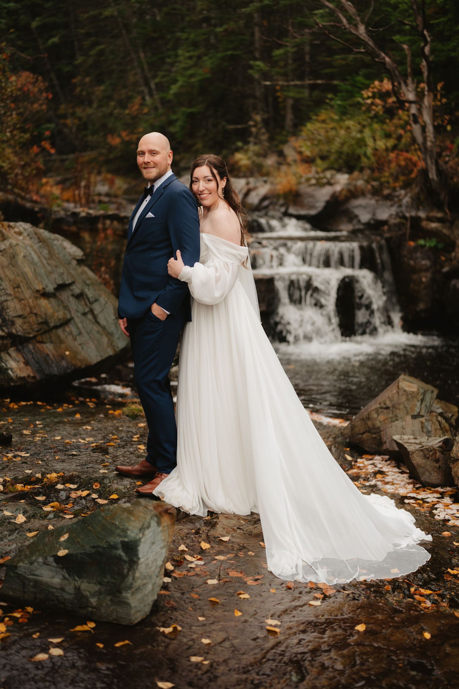 A couple in wedding attire poses by a cascading waterfall surrounded by autumn foliage and fallen leaves.