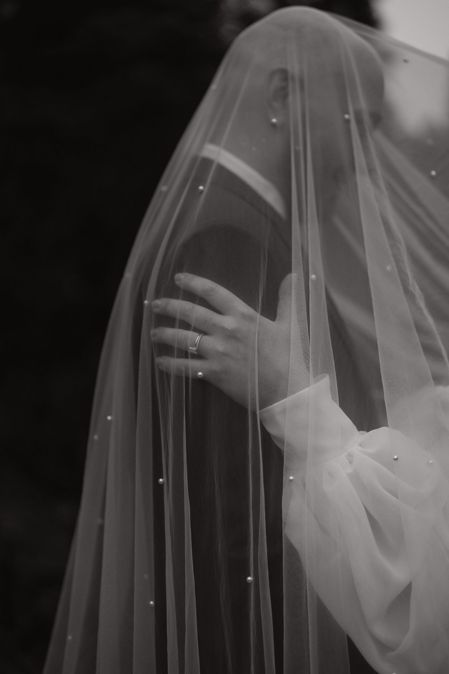 Black and white close-up photo of a delicate bridal veil with scattered pearl details flowing in soft light.