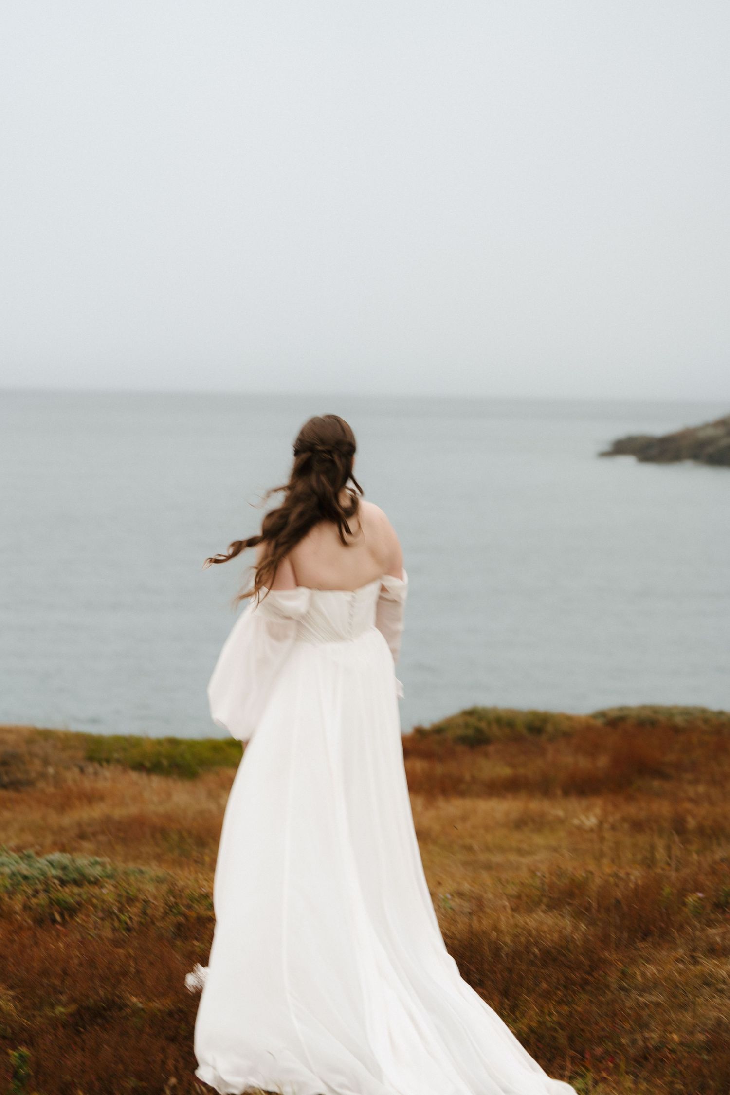 A person in a flowing white dress walks along a windswept coastal cliff with ocean views in the background.