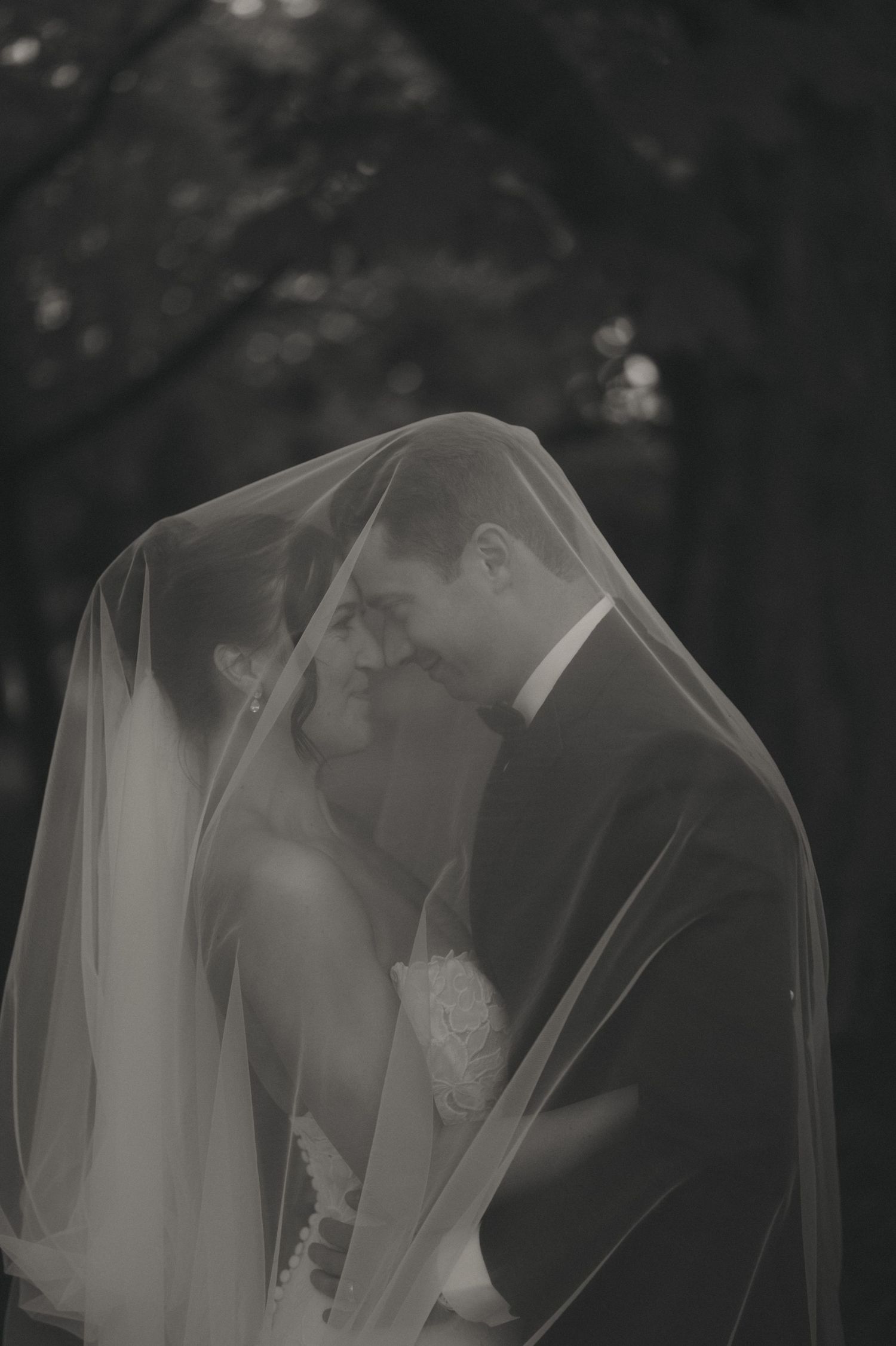 Black and white photo of bride and groom sharing a romantic moment under a flowing wedding veil outdoors.
