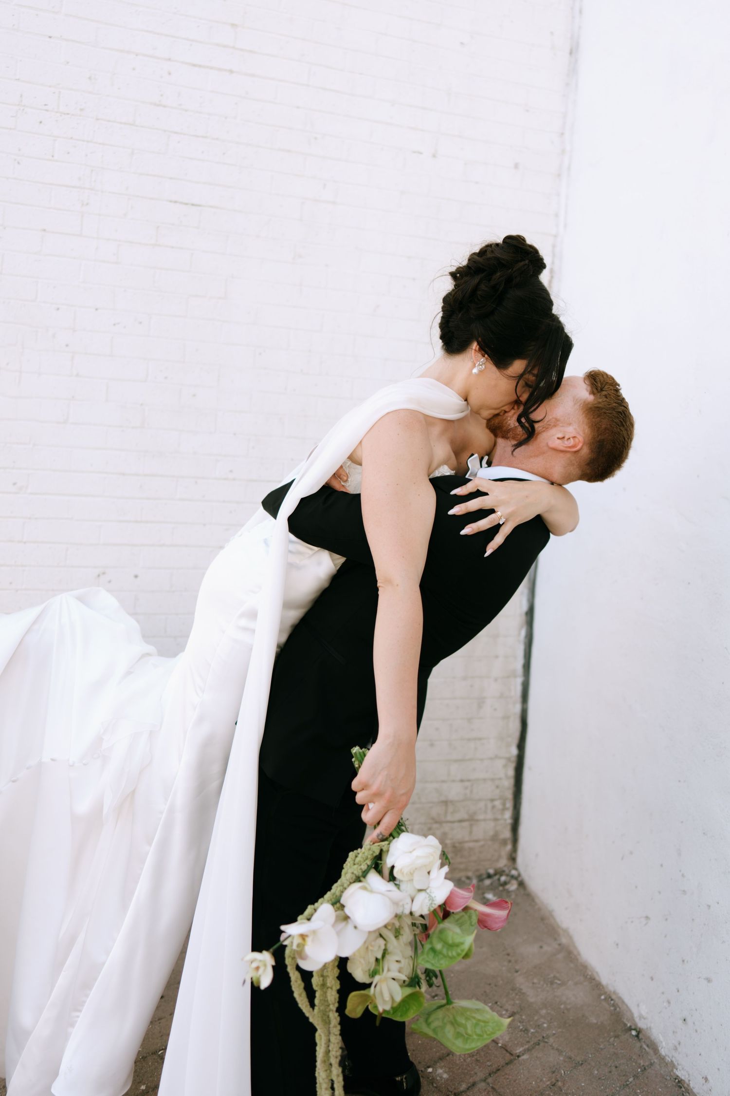 Bride in white gown kissing groom in black suit against white brick wall, holding white floral bouquet.