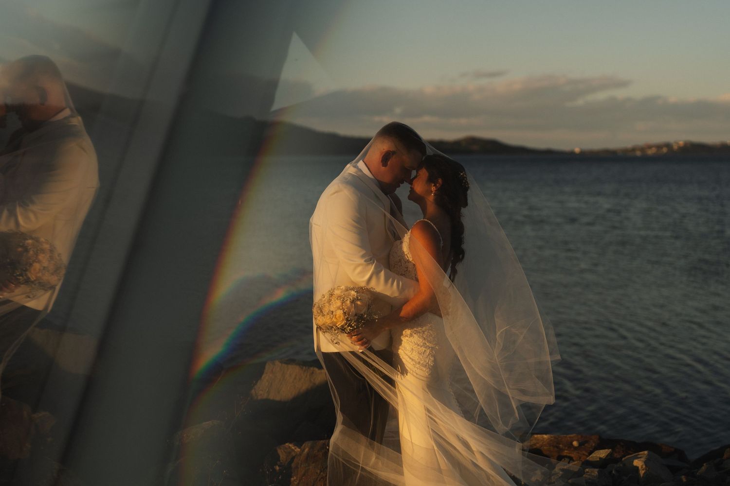 Romantic couple embraces during golden hour sunset by the ocean while wearing wedding attire.