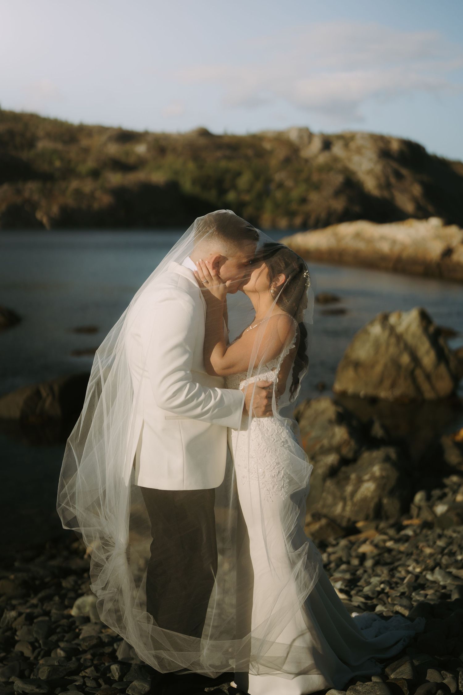 A romantic couple shares an intimate moment by the rocky coastline during a sunset wedding photoshoot.