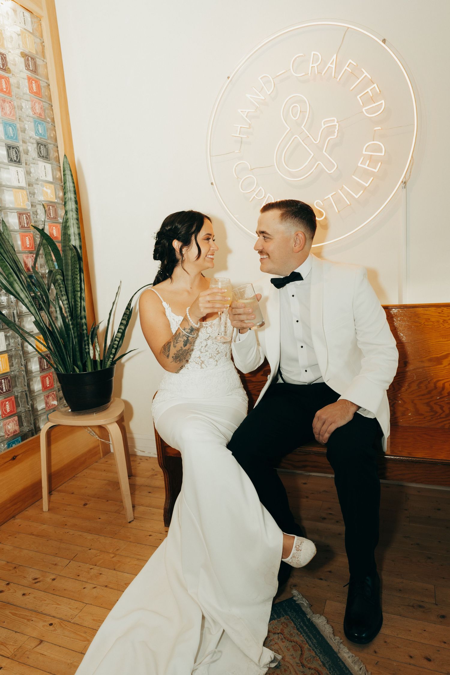 A couple in wedding attire share a romantic moment on a wooden floor with neon signs glowing on the wall behind them.