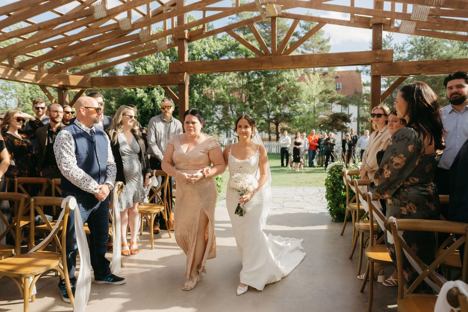 A bride walks down the aisle during an outdoor wedding ceremony under a rustic wooden pavilion with guests seated on both sides.