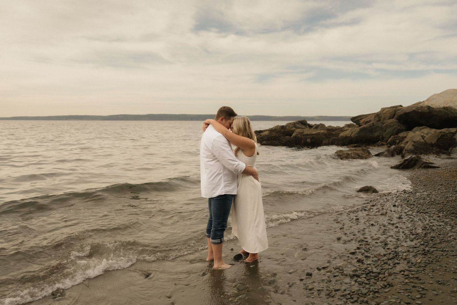 A couple embraces while standing in shallow ocean water at sunset along a rocky beach shore.